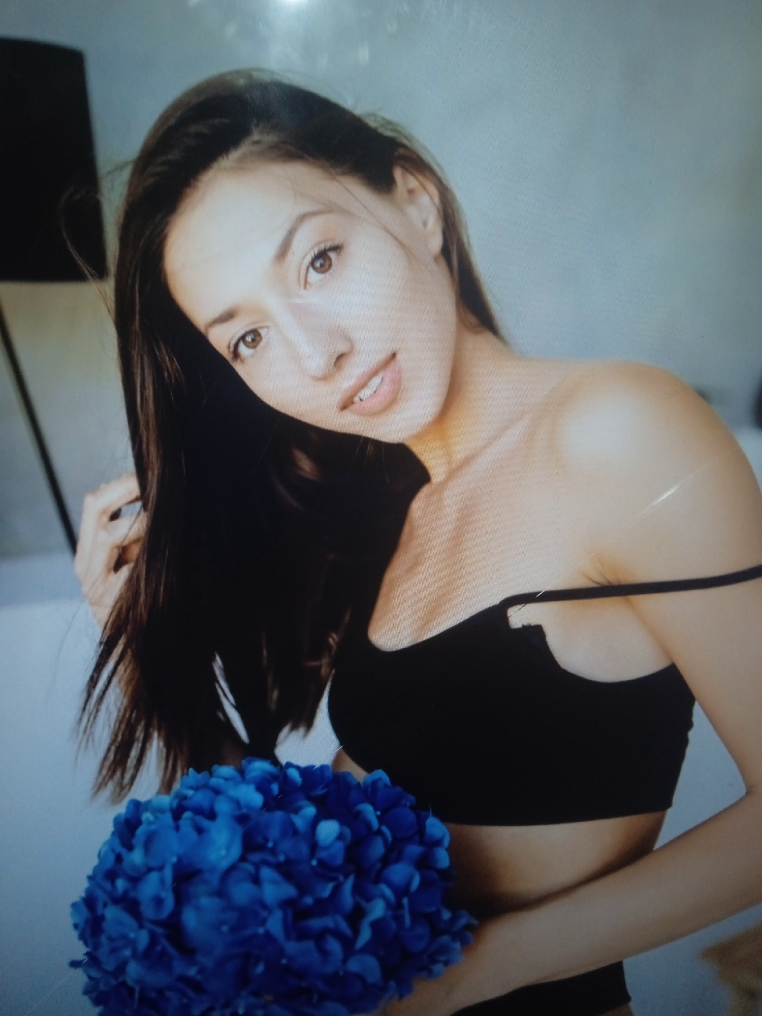 A young woman with long, dark hair is positioned in the center of the frame, looking towards the viewer. She is wearing a black, strappy top, with one strap hanging off her shoulder, revealing her upper chest and shoulder. Her left hand is raised and gently touching her hair. In front of her, a large bouquet of vibrant blue hydrangeas is held. The background is a soft, light blue, suggesting an indoor setting. The overall lighting is soft and diffused, highlighting the woman and the flowers. There is no discernible location within the city of Chernivtsi, Ukraine.