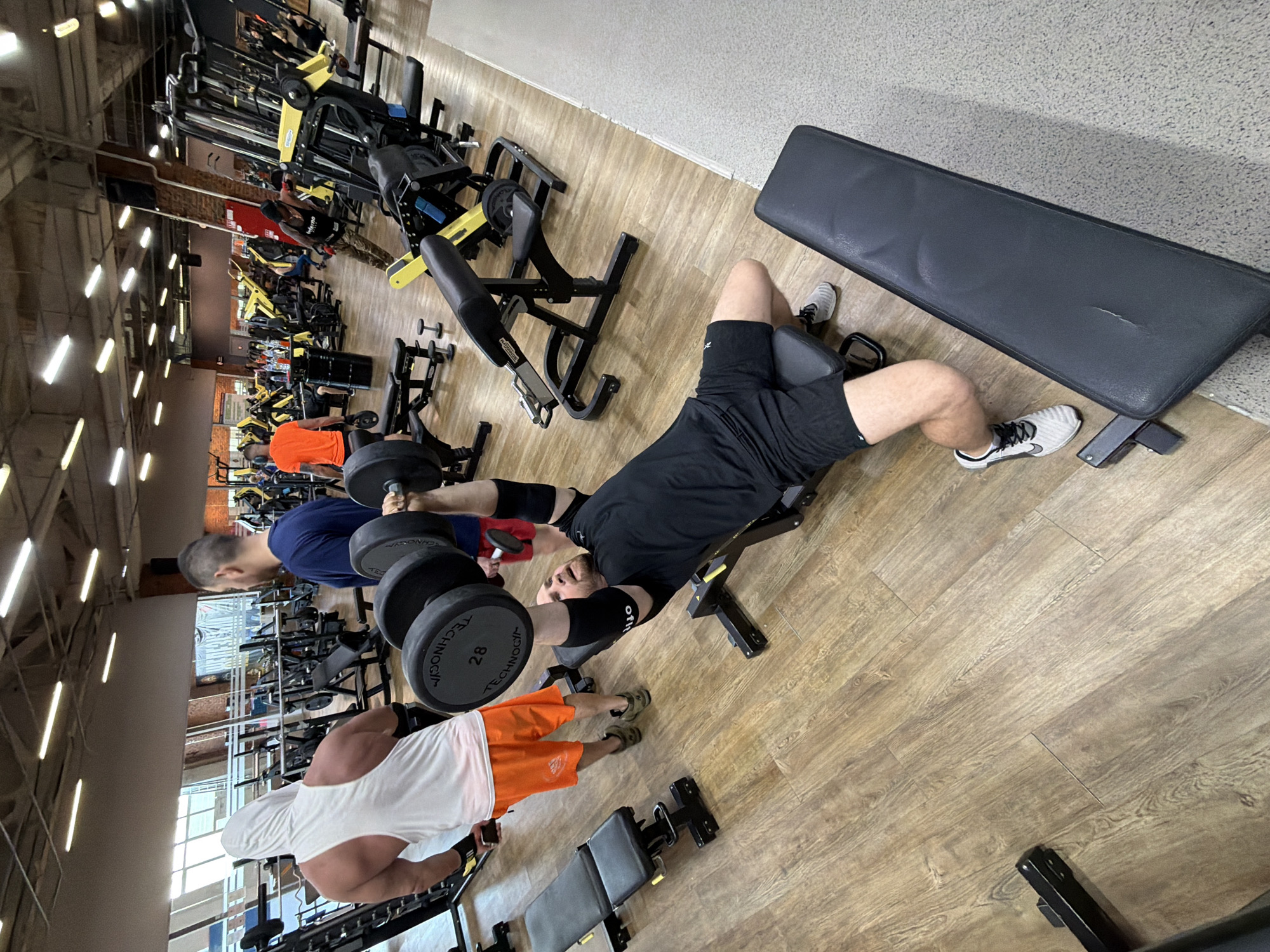 The image shows a gym interior with several people engaged in weightlifting. In the foreground, a man in black shorts and shirt is lying on a weight bench, lifting dumbbells. Another man wearing a blue shirt is also lifting dumbbells nearby. A third person in a white shirt and orange shorts is seen at the bottom of the frame. Gym equipment is visible throughout the scene. The location is most likely Chaikovsky, Russia.