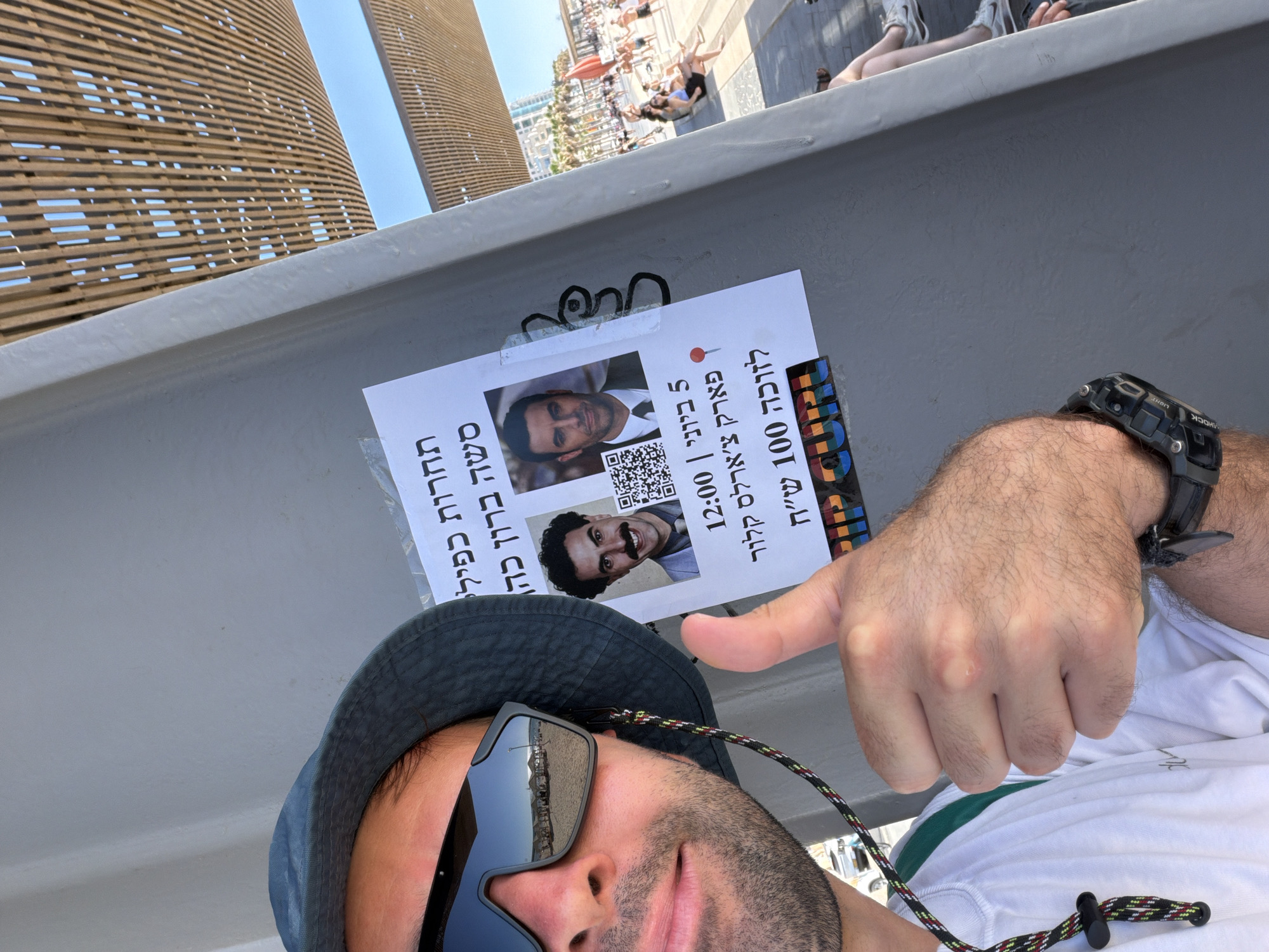 The image is a selfie-style shot taken outdoors on a bright, sunny day in Tel Aviv, Israel. In the foreground, a man, partially visible, wears a dark bucket hat, sunglasses reflecting the surroundings, a white t-shirt, and a black G-Shock watch on his wrist. His right hand, with a hairy arm, is raised with the thumb pointing upwards, gesturing towards a flyer.
The flyer, taped to a grey horizontal beam, is written in Hebrew and advertises a "Sacha Baron Cohen look-alike contest" ("תחרות כפילי סשה ברון כהן"). It features two photographs of men, one clearly resembling Borat, a character played by Sacha Baron Cohen, along with two QR codes. The text further indicates a prize of "100 NIS for the winner" ("לזוכה 100 ש"ח") and states the event is scheduled for "June 5 | 12:00" ("5 ביוני | 12:00") at "Charles Clore Park" ("פארק צ'ארלס קלור").
The background reveals a lively outdoor scene under a clear blue sky. Numerous people are visible, mostly lounging and relaxing in a public space, possibly a park or promenade, consistent with Charles Clore Park's location. Modern buildings with distinct horizontal wooden slat facades line the background, adding to the urban setting. The man's thumbs-up gesture suggests enthusiasm or endorsement of the advertised event.