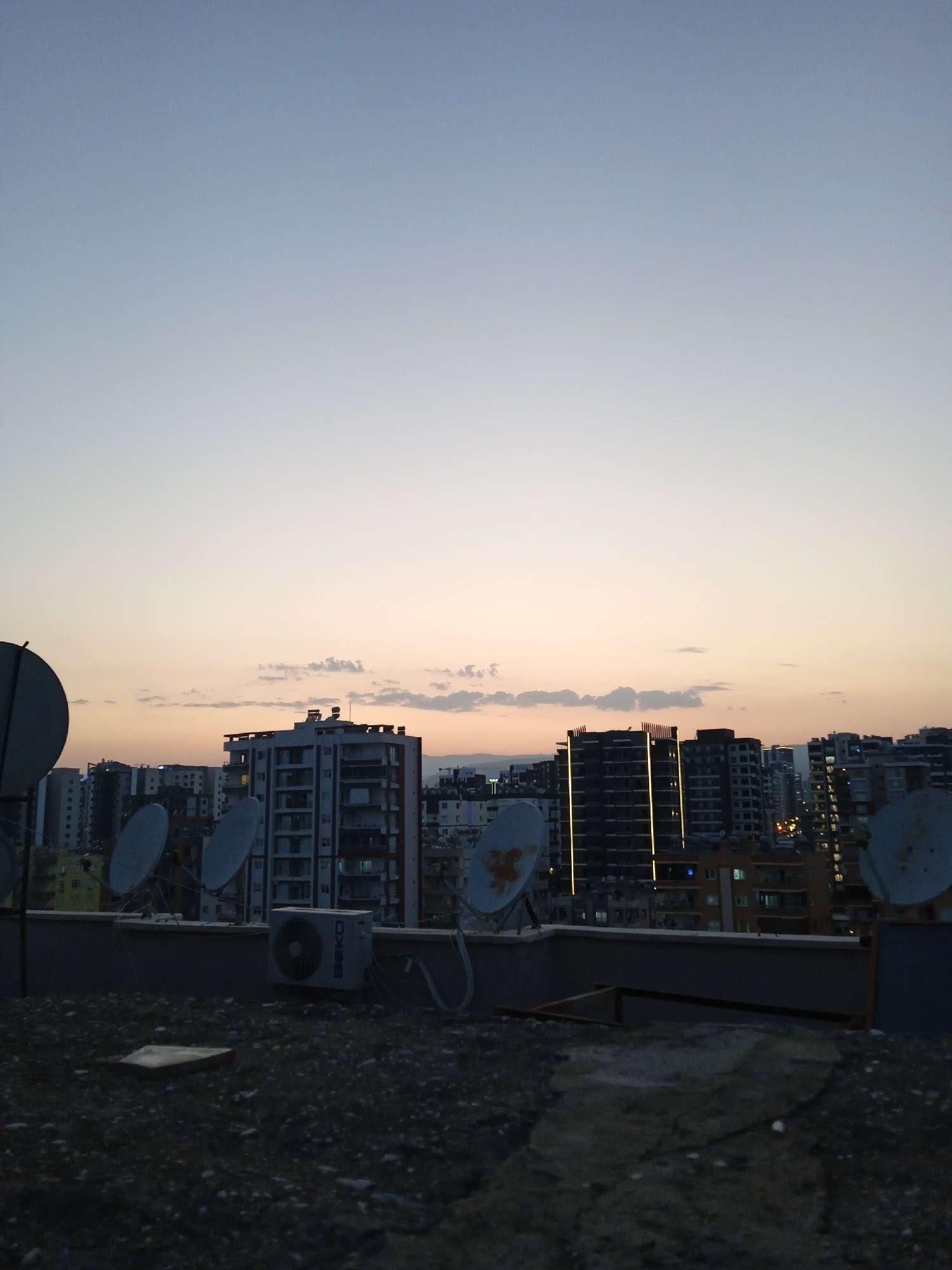 The image depicts a rooftop view of a city skyline at dusk, with buildings and satellite dishes visible. The sky is a gradient of pale blue and orange, with a few wispy clouds. In the foreground, a dark, pebbled rooftop is visible, with a square, flat object in the lower left corner. Several satellite dishes, some appearing weathered, are positioned on the rooftop. Behind the rooftop, a series of multi-story buildings dominate the cityscape. The buildings vary in architectural style and color, with some featuring prominent windows and balconies. Lights are visible in some of the buildings, indicating evening or night. The overall impression is one of an urban environment transitioning from day to night. The provided hint suggests the location is Mezitli, Turkey.
