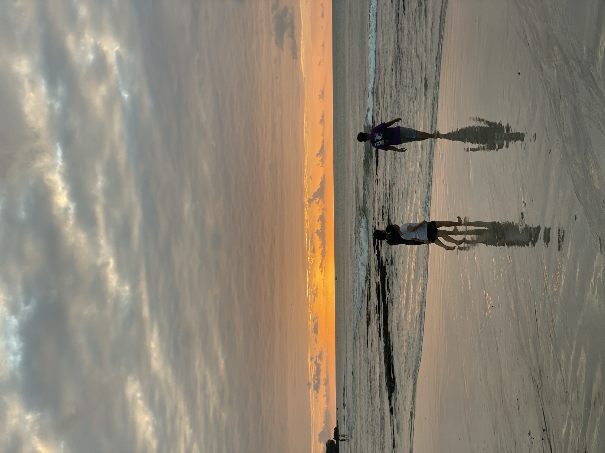 The image depicts a serene beach scene in Kuta Selatan, Indonesia, during a sunset. The sky is filled with a gradient of colors, from soft oranges and yellows near the horizon to muted grays and blues overhead, with scattered clouds. The sun is low, casting a warm glow across the water and wet sand. Gentle waves lap at the shore, creating a reflective surface on the beach.
In the foreground, three figures are visible walking along the water's edge. To the left, two individuals, likely a child and an adult, are walking together. The child, wearing a white top and dark shorts, is slightly ahead, while the adult, in a dark top, walks behind. To the right, a third person, wearing a purple t-shirt and gray shorts, walks away from the camera towards the ocean. Their reflections are clearly visible on the wet sand, adding depth to the scene.
Further out in the water, on the left side of the frame, a small group of people can be seen walking in the shallow water near some dark rocks. A distant boat is also visible on the horizon. The overall atmosphere is peaceful and tranquil, characteristic of a sunset at a tropical beach.