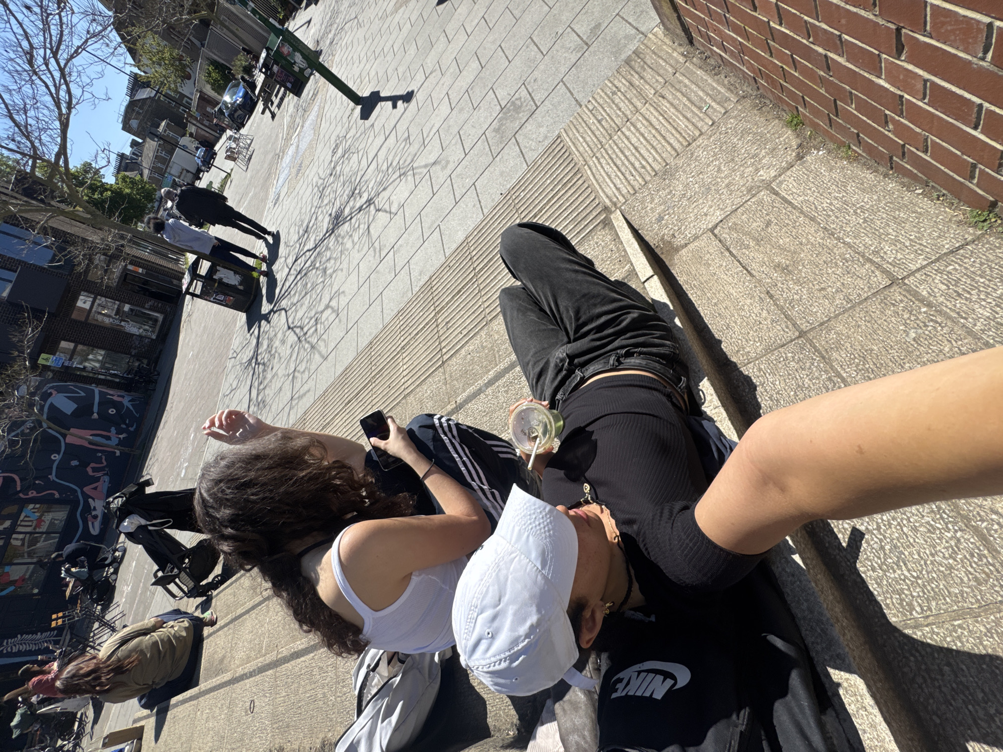 The scene is an outdoor shot, presumably taken by someone who is part of the group. Three people are visible, two closer to the camera and one further away. One person in a white tank top and curly brown hair sits near the camera, looking at a phone in her hand. Beside her, another person is lying down, wearing a white cap and a black Nike t-shirt. They appear to be holding a drink. The photographer's arm is partially visible on the right.
In the background, other pedestrians are walking along the sidewalk. Buildings of varied heights surround the area. The ground is paved with stone tiles. The brick wall can be seen on the right side of the picture.
Based on architectural styles and street layout, the scene could be located in the city of London, United Kingdom.