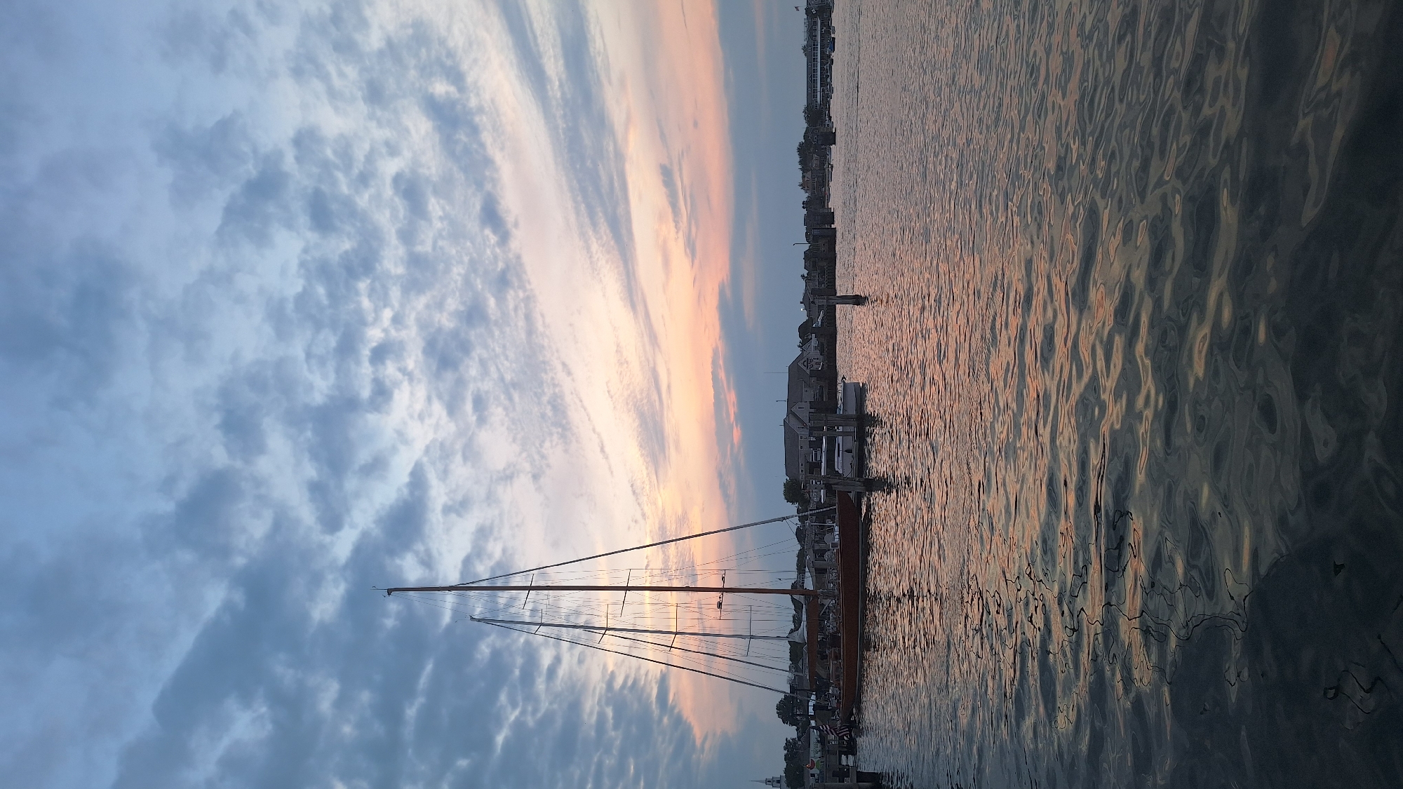 A panoramic view captures the sunset over Nantucket Harbor, USA, with a large sailboat prominently docked in the foreground. The sailboat, with its tall masts and wooden hull, rests on calm water that reflects the soft hues of the twilight sky. Several smaller boats are also moored nearby, alongside a pier. In the background, the Nantucket coastline is dotted with buildings and structures, including what appears to be a church steeple, bathed in the warm glow of the setting sun. The sky is a canvas of soft blues and pinks, streaked with clouds that are beginning to darken as night approaches. The overall atmosphere is serene and tranquil, conveying a peaceful end to the day.
