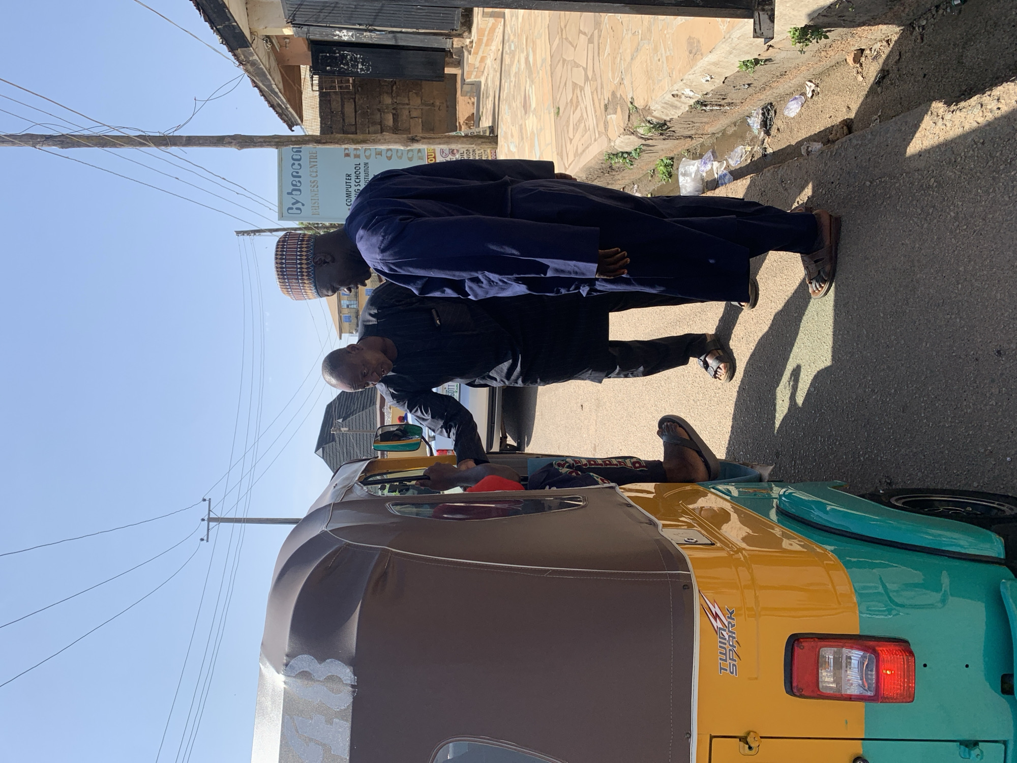 The image shows two men standing near a yellow and green "Twin Spark" tricycle in Jos, Nigeria. One man wears a dark suit and the other a blue robe and a patterned hat. They are likely interacting with the tricycle driver. The environment appears to be a street with buildings and a "Cybercon Business Centre" sign visible. There are power lines overhead. The ground is paved.