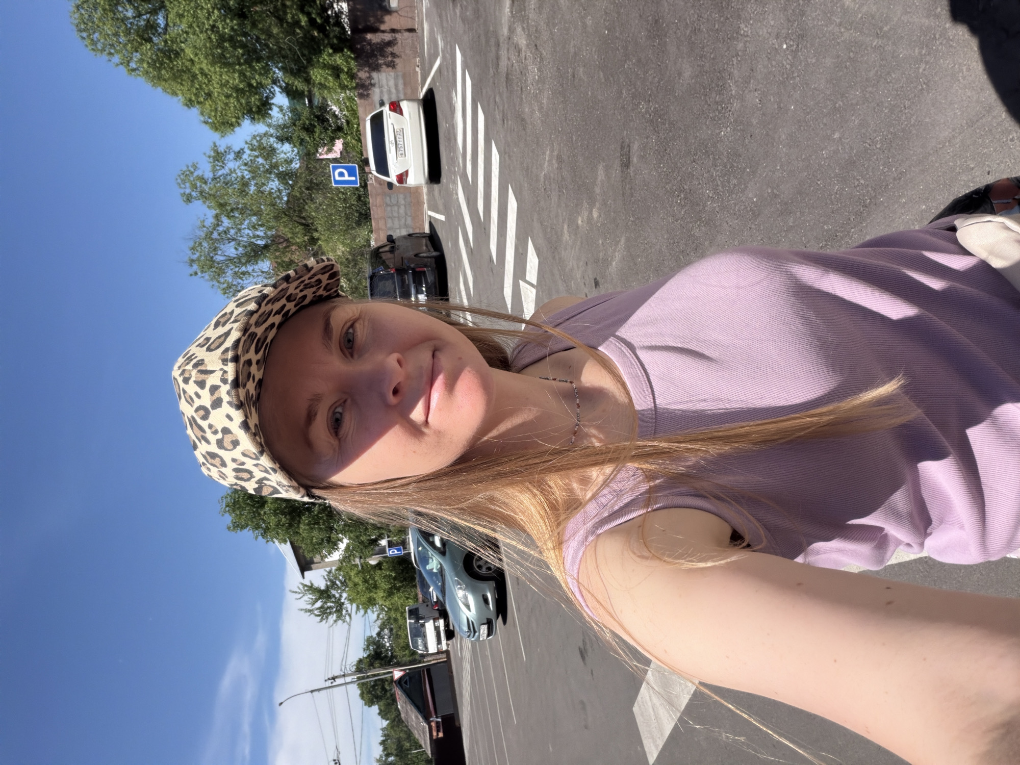 A woman wearing a leopard-print baseball cap and a purple tank top is taking a selfie outdoors. She is standing in a paved parking lot with white pedestrian crossing lines. Several cars are parked in the lot, including a white sedan, a black van, and a light blue sedan. In the background, there are trees and buildings. A blue parking sign with a white 'P' is visible. The scene appears to be in a city, possibly Khabarovsk, Russia, given the license plates visible on the cars. The weather is sunny, with a clear blue sky.