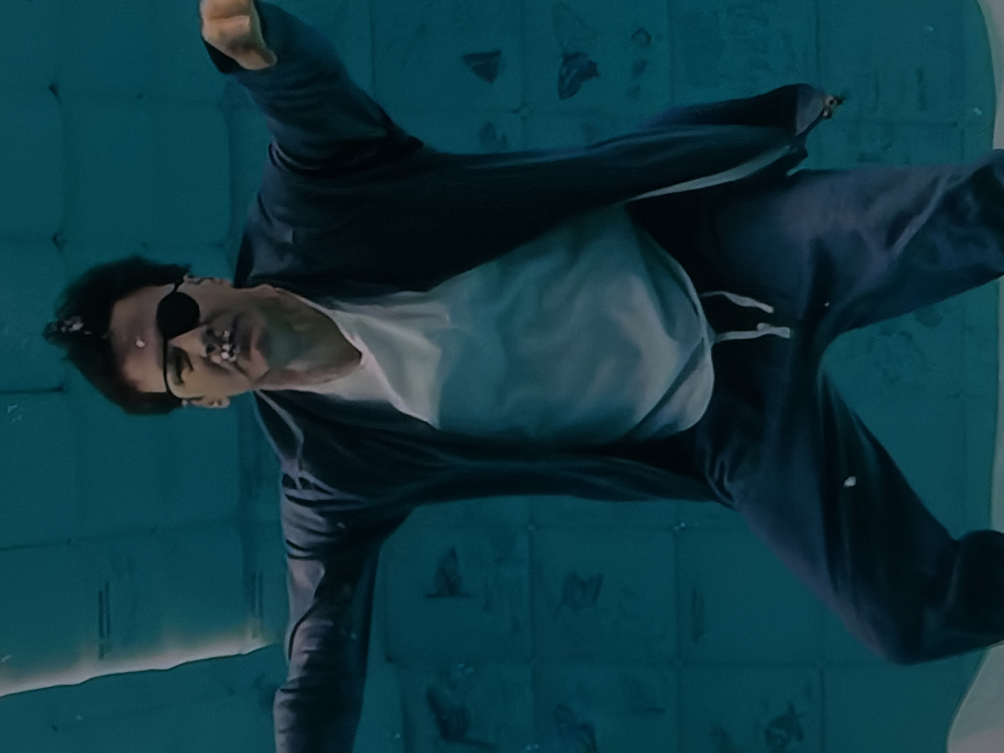 A person is depicted submerged underwater in what appears to be a swimming pool or a large water tank in Sofiivska Borshchahivka, Ukraine. The subject, likely male, is wearing a dark jacket or tracksuit top, a white t-shirt, and dark trousers. They are also wearing dark sunglasses. The individual is suspended at an angle in the water, with their head slightly lower than their feet, suggesting they are either floating or gently sinking. Small bubbles are visible around their mouth and face, indicating exhalation or air escaping.
The underwater environment is characterized by a consistent teal-blue hue, with visible square patterns that resemble pool tiles on the background wall. There are some faint, irregular shapes or reflections on these tiles. The lighting in the scene is diffused, illuminating the person and the water. The overall impression is one of calm and stillness. No visible text or specific time of day is discernible from the image.