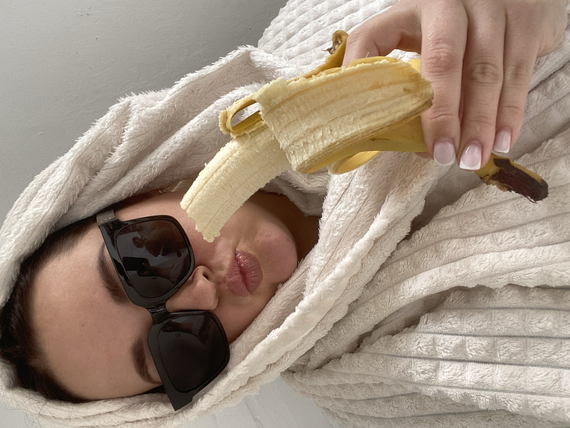 A woman wearing black sunglasses and wrapped in a white terry cloth robe is holding a partly peeled banana in front of her face. Her lips are pursed in a kissing pose. The woman's fingernails are manicured with a white-tipped French style. The background appears to be a neutral-toned wall. There is no visible evidence to confirm the location as Sochi, Russia.