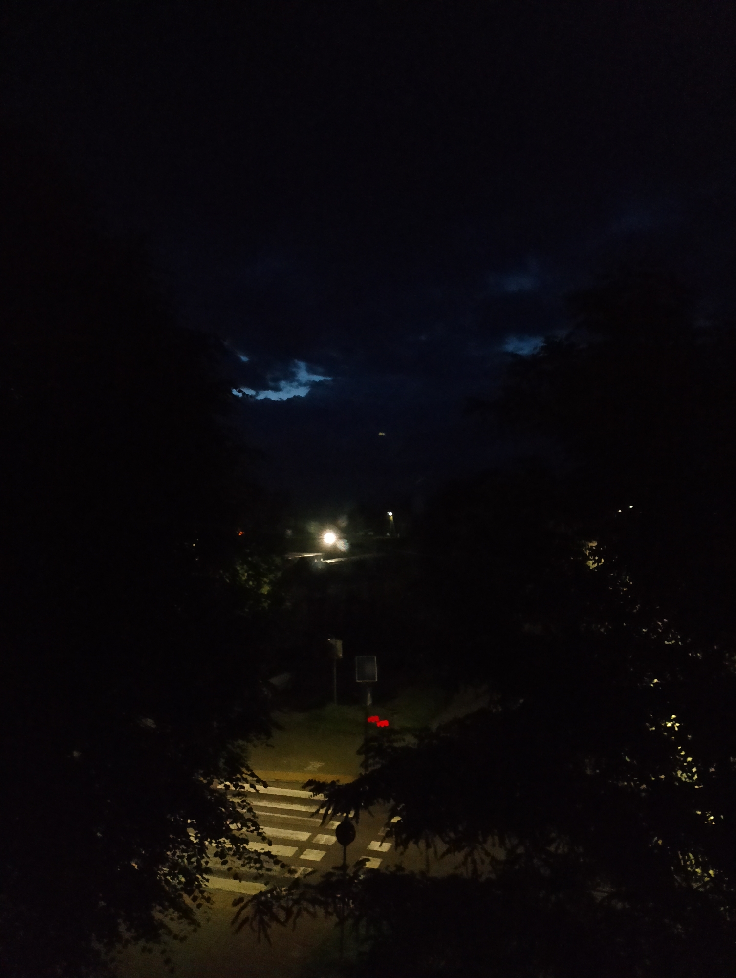 A nighttime, high-angle view shows a dark, cloudy sky with patches of lighter blue visible. In the foreground, silhouetted trees frame the scene. Below, a street scene unfolds, illuminated by artificial lights. A crosswalk with bright white stripes is visible on the asphalt. A traffic light with red signals is mounted on a pole. In the distance, streetlights cast a glow on the road and surrounding foliage. The overall impression is of a quiet, urban night in Kolno, Poland.