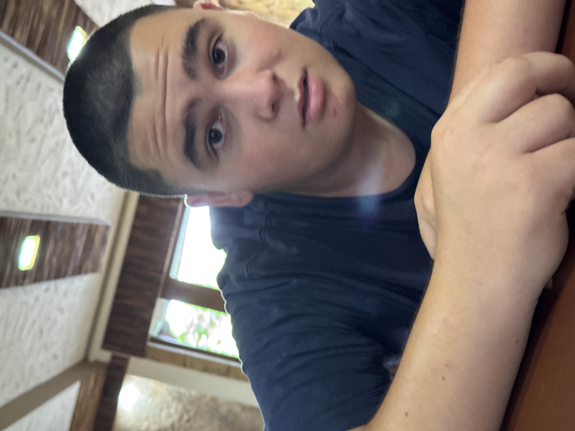 A young Caucasian male with a buzz cut is pictured from the chest up, leaning forward with his arms crossed in front of him. He is wearing a dark blue t-shirt. He has a fair complexion, dark eyes, and appears to be looking directly at the camera with a neutral expression. The background shows a ceiling with wooden beams and recessed lighting, and a window with greenery visible outside. The setting appears to be indoors, possibly a restaurant or a similar public space. The composition is a close-up, with the subject occupying most of the frame. The lighting is natural and coming from the front, creating some slight glare on his forehead and t-shirt.