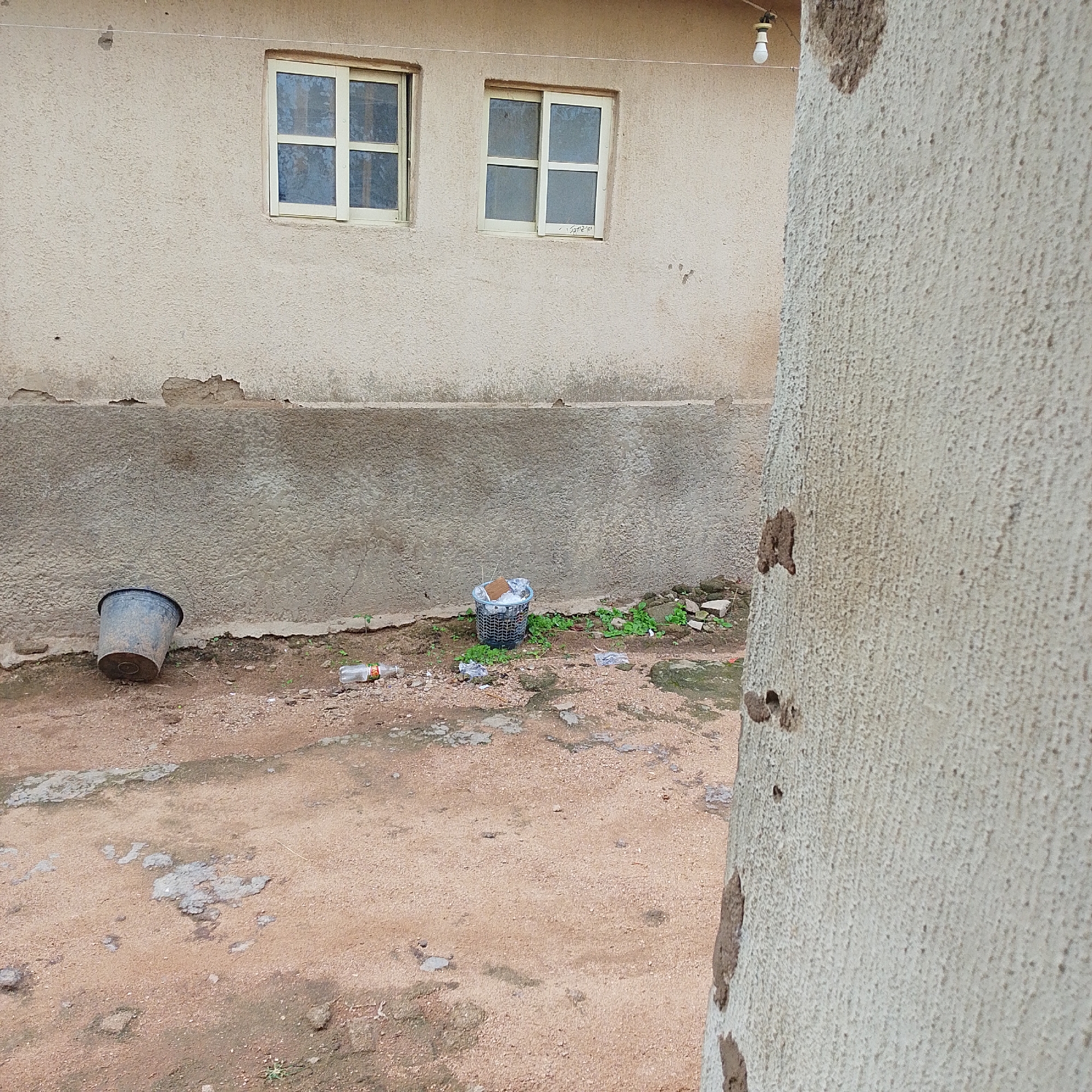 The image shows an outdoor scene in Jalingo, Nigeria. The foreground is dominated by a rough, sandy ground with patches of concrete and some scattered green vegetation. In the midground, a dark gray plastic bucket is overturned on the ground to the left, and a small wire mesh basket filled with debris is situated to the right. Scattered on the ground are pieces of trash, including a plastic bottle. Behind these objects, a building with a textured, light-colored facade features two windows with white frames and divided panes. The lower part of the building's wall has a rough, concrete band. The background is out of focus, but appears to be more of the same building. A single light bulb hangs from a wire on the upper right side of the frame. The right edge of the image is filled by a close-up view of a textured, light-colored wall, possibly part of another structure, with several dark, irregular patches on its surface.