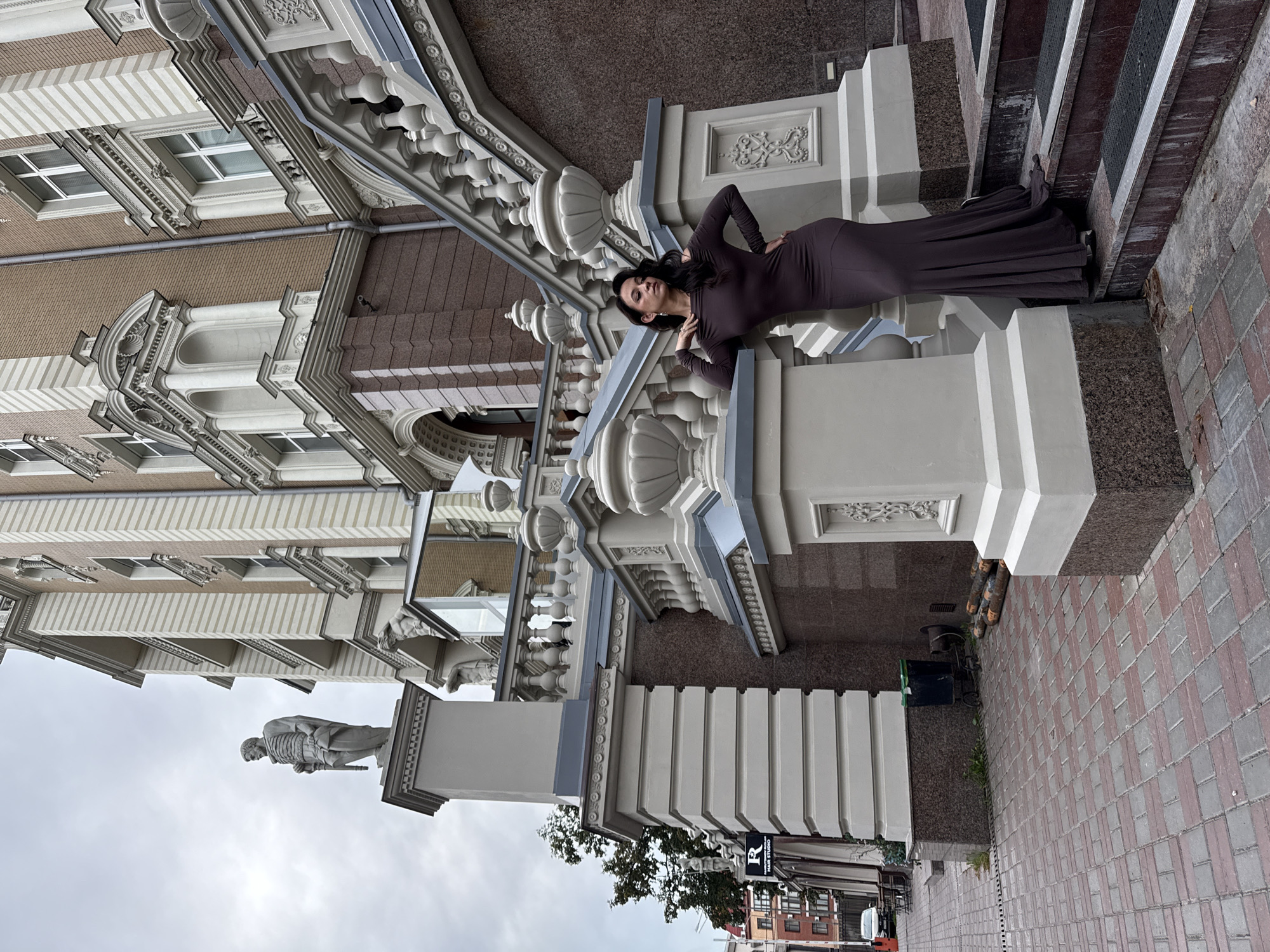 The image depicts a woman standing on a grand, ornate staircase outside a building. The woman is wearing a long, dark brown, form-fitting dress that flares out at the bottom. She has dark hair and is posing with one hand on a railing and the other on her hip. The staircase is made of stone with decorative railings and balustrades. The building behind her is tall and features classical architectural elements with brick and stone accents. A statue of a man in historical attire stands prominently on a pedestal to the left of the staircase, near the top of a large column. Below the staircase, on the ground level, a paved sidewalk with a brick pattern is visible. A sign for "R Fashion House Hair Studio" is attached to the side of a pillar near the sidewalk. The overall scene suggests an urban setting, likely in a historically significant area. This building is part of the "AURA Business Park" in Kazan, Russia.