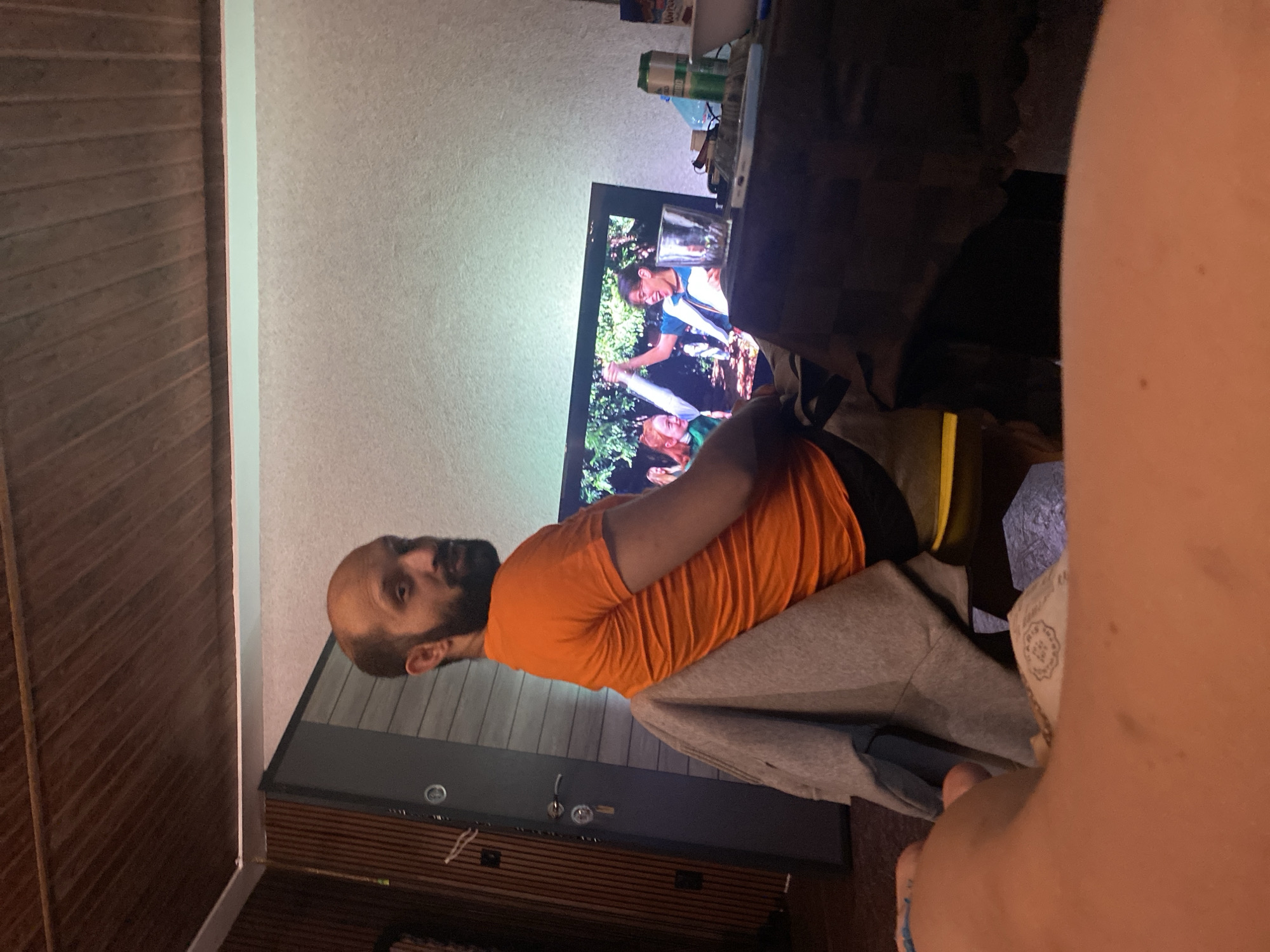 The image captures an indoor scene, likely a living room, from a slightly elevated, first-person perspective, with the photographer's bare arm and shoulder visible in the right foreground. A blue string or bracelet is on the arm, and part of a dollar bill is also visible.
The main subject is a man with a bald head and a beard, wearing an orange t-shirt and grey sweatpants. He is seated on a grey couch or armchair, his body angled towards a television screen, and he appears to be watching the content displayed.
The television, positioned centrally, shows what seems to be a show or movie. On the screen, two individuals are visible in an outdoor setting with lush greenery. One is a young woman with dark hair and glasses, wearing a white and blue top, depicted in a moment of apparent joy or excitement. Another person with red hair is partially visible at the bottom edge of the screen. A clear glass is resting on top of the television.
Behind the television, on a shelf or surface, various items are scattered, including several cans (one possibly a "Whiteclaw" product) and other unidentifiable objects. The room features a light-colored, textured wall on the right side and a dark wooden slatted wall or paneling on the left, separated by a thin vertical white strip. Below the TV, there's a dark piece of furniture with some circular details. The lighting suggests it might be evening or night, with the television screen illuminating parts of the scene. The overall impression is one of casual home entertainment.