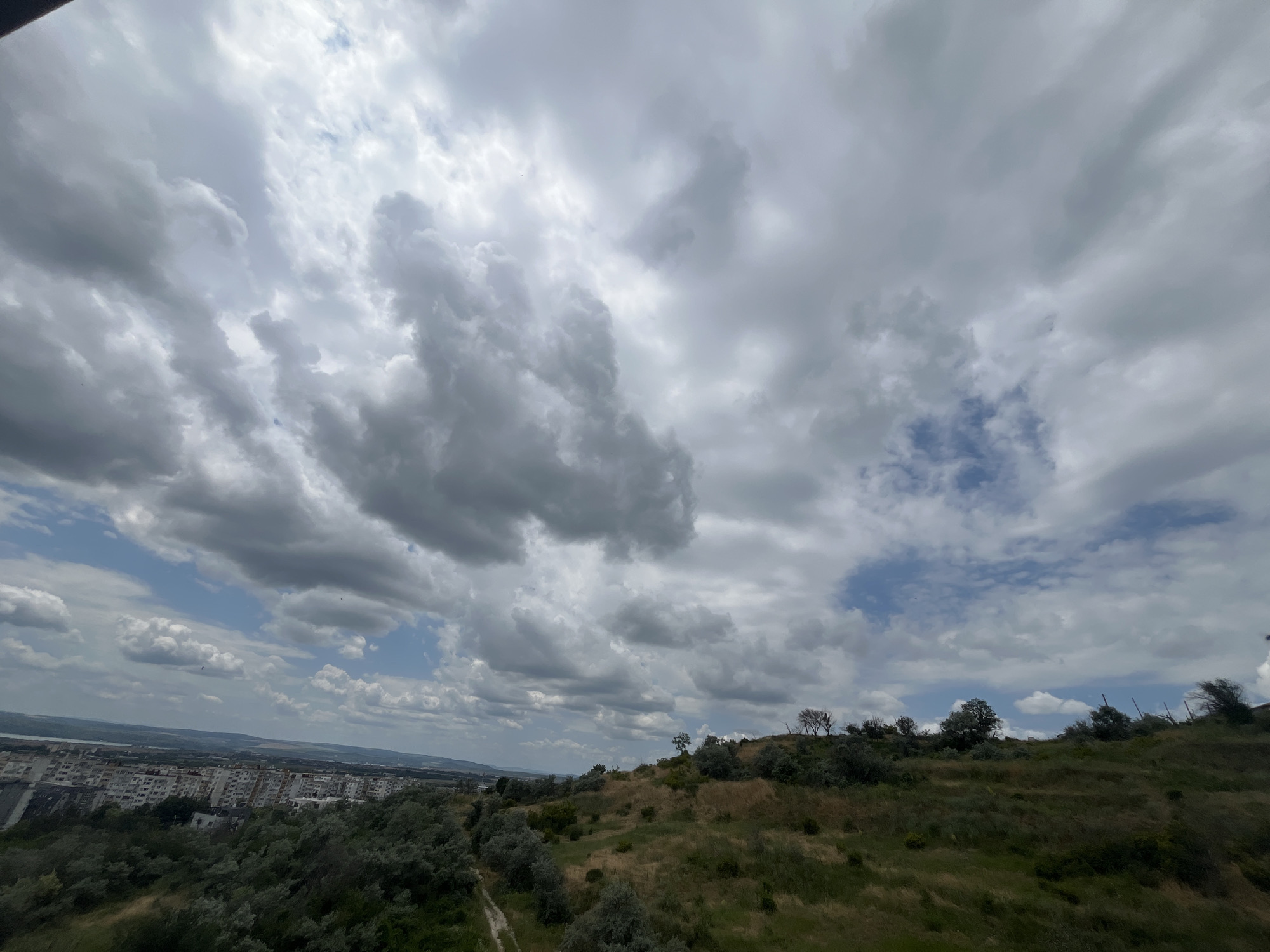 The image depicts a wide-angle view of a hilly landscape overlooking a city under a cloudy sky. In the foreground, a grassy slope with scattered trees and shrubs dominates the right side of the frame, with a dirt path winding through it. The middle ground features a dense cluster of trees and vegetation, with some buildings of a city partially visible through them on the left. The city itself, with its blocky apartment buildings, extends towards the horizon on the left. The background shows a body of water, likely the sea, and the distant horizon. The sky is filled with dramatic, heavy clouds, with patches of blue sky visible in between. The overall impression is a semi-urban environment with significant natural elements, possibly taken from a viewpoint on a hill. This scene is consistent with viewpoints found in the Varna city quarters of Briz or Chaika, where hilly terrain meets the urban landscape and offers views towards the sea and the city. There are no people or specific actions identifiable in the image.