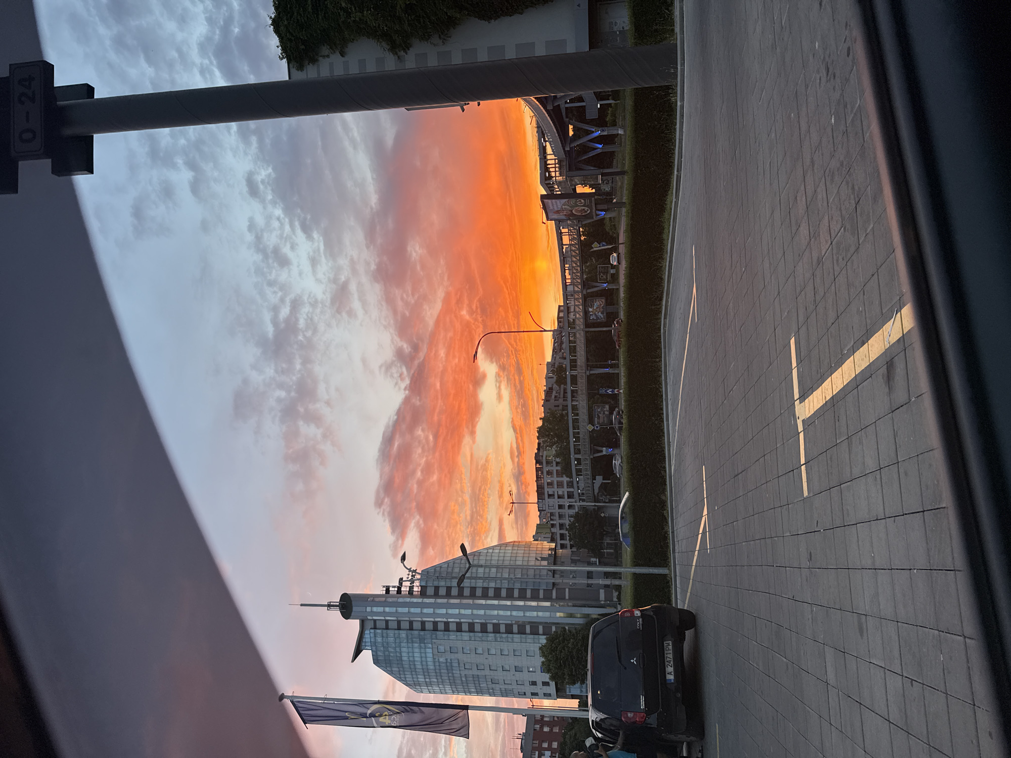 The image captures a vibrant sunset over the city of Burgas, Bulgaria, viewed from a parking lot. In the foreground, a dark-colored hatchback car, a Mitsubishi Colt, is parked in a paved lot marked with yellow lines. The car's license plate reads "A 2471 PM". A tall, dark gray lamppost with a sign indicating "0-24" stands to the right of the frame.
Dominating the mid-ground is a modern, multi-story glass-facade building with a distinctive curved design, topped with a spire. Next to it, a dark blue flag with a yellow emblem is displayed on a flagpole. Across the street, a pedestrian overpass with illuminated blue supports spans the road, with billboards visible on its sides. In the background, a cluster of residential buildings and other structures are silhouetted against the colorful sky.
The sky itself is a dramatic canvas of fiery oranges, reds, and soft pinks, with scattered clouds catching the last rays of the setting sun. The overall scene suggests an urban environment at dusk, with the natural beauty of the sunset juxtaposed against man-made structures. The photograph appears to have been taken from inside a vehicle, given the dark framing at the bottom of the image.