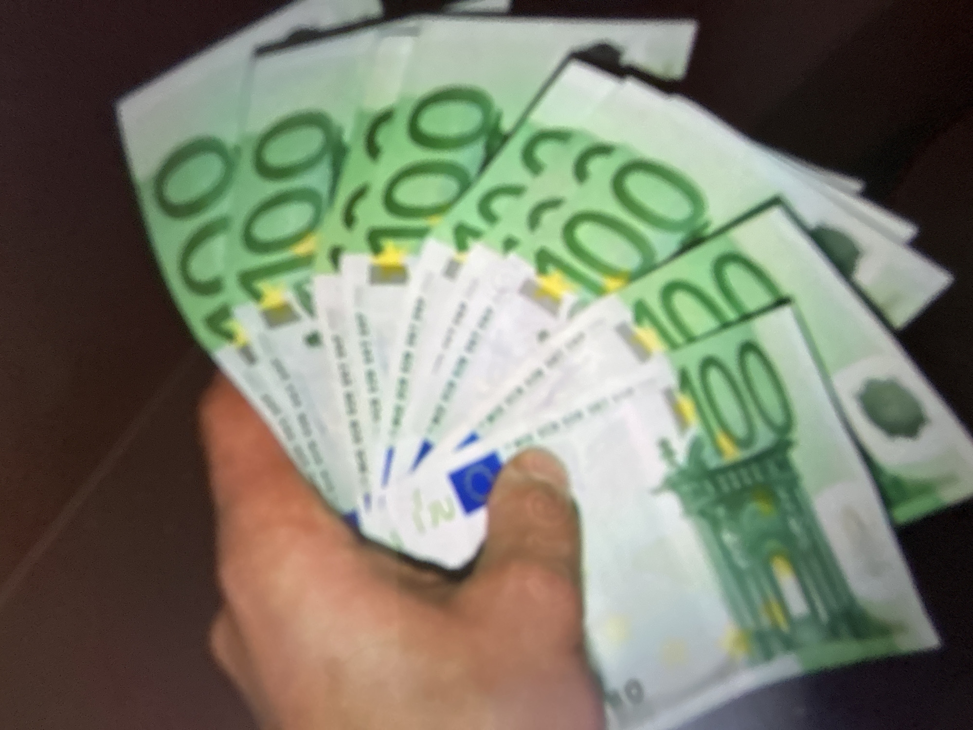 The media file is a close-up, slightly blurred photograph of a person's hand holding a fan of 100 Euro banknotes. The banknotes are predominantly green and feature the numeral "100" prominently on them, along with architectural imagery. The hand appears to be that of an adult. The background is dark and out of focus, suggesting an indoor setting or a dimly lit environment. There are no discernible environmental cues indicating the time of day or weather conditions. The image primarily focuses on the money and the hand, with no other people or significant background elements visible. The overall impression is one of wealth or financial transaction. No text other than the denomination "100" on the banknotes is clearly legible. The location context is provided as Gelendzhik, Russia.