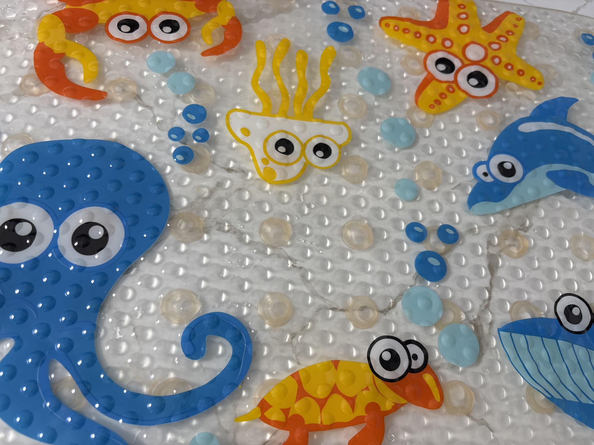 The image shows a close-up view of a non-slip bath mat decorated with cartoon sea creatures. There is a blue octopus, a yellow and orange crab, a yellow and orange starfish, a white and yellow jellyfish, a blue dolphin, and a yellow and orange turtle. Blue and light blue bubble shapes are interspersed among the creatures. The mat itself has a white, slightly marbled surface with many clear, small, raised suction cups. The image does not appear to be related to Chaikovsky, Russia.