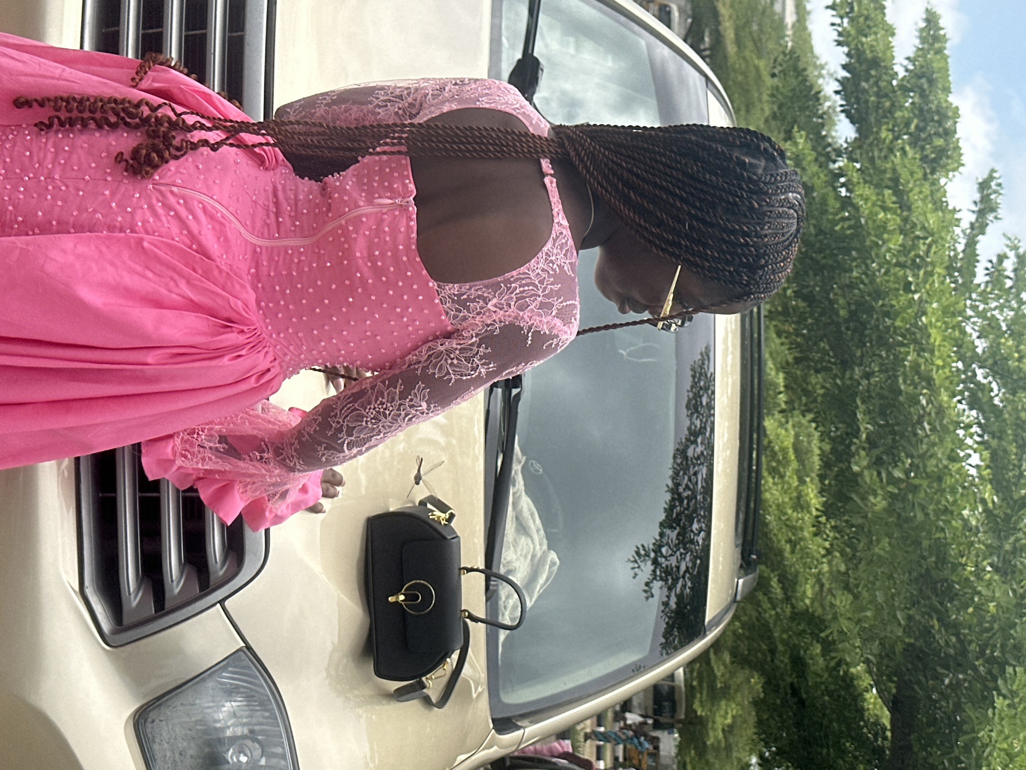 A young woman with braided hair, wearing a pink sequined dress and gold-rimmed glasses, stands with her back to the camera, leaning on the hood of a gold SUV parked outdoors. A black handbag rests on the hood. The scene is set against a backdrop of lush green trees and a cloudy sky. The location is identified as Itoga, Nigeria.