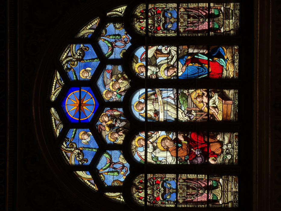 A multi-paneled stained glass window, set within a dark, arched architectural frame, depicts a religious scene. This window is located in a church or cathedral in Paris, France.
The central lower section features the Nativity. To the left, a bearded male figure (Joseph), adorned with a golden halo, kneels on one knee, hands clasped in prayer, and gazes towards the center. He wears a red tunic and a blue garment. In the immediate center, an infant (Baby Jesus), also with a golden halo, lies swaddled in white cloth within a wooden manger filled with hay. To the right, a female figure (Mary), wearing a blue gown, red underskirt, and a white head covering, kneels with a golden halo, looking reverently at the infant. White flowers are visible on the ground near Joseph.
Above the central figures, a series of angels with white robes and golden halos are depicted across several arched panes. Some angels have hands clasped in prayer, while others hold scrolls or banners, one with visible text. The upper portion of the window consists of hexagonal panes, predominantly featuring a blue honeycomb pattern. A prominent central hexagonal pane displays an eight-pointed orange star on a deep blue background, framed by a gold band. Surrounding this are other hexagonal panes containing additional angelic figures and decorative elements.
Vertical, narrower panels flank the main Nativity scene on both sides. These panels showcase intricate patterns in red, blue, and gold, including symbols resembling a chalice and rosary beads on the left-most panel. The entire window is rendered in vibrant hues of blue, red, gold, yellow, and green glass, with varying textures.