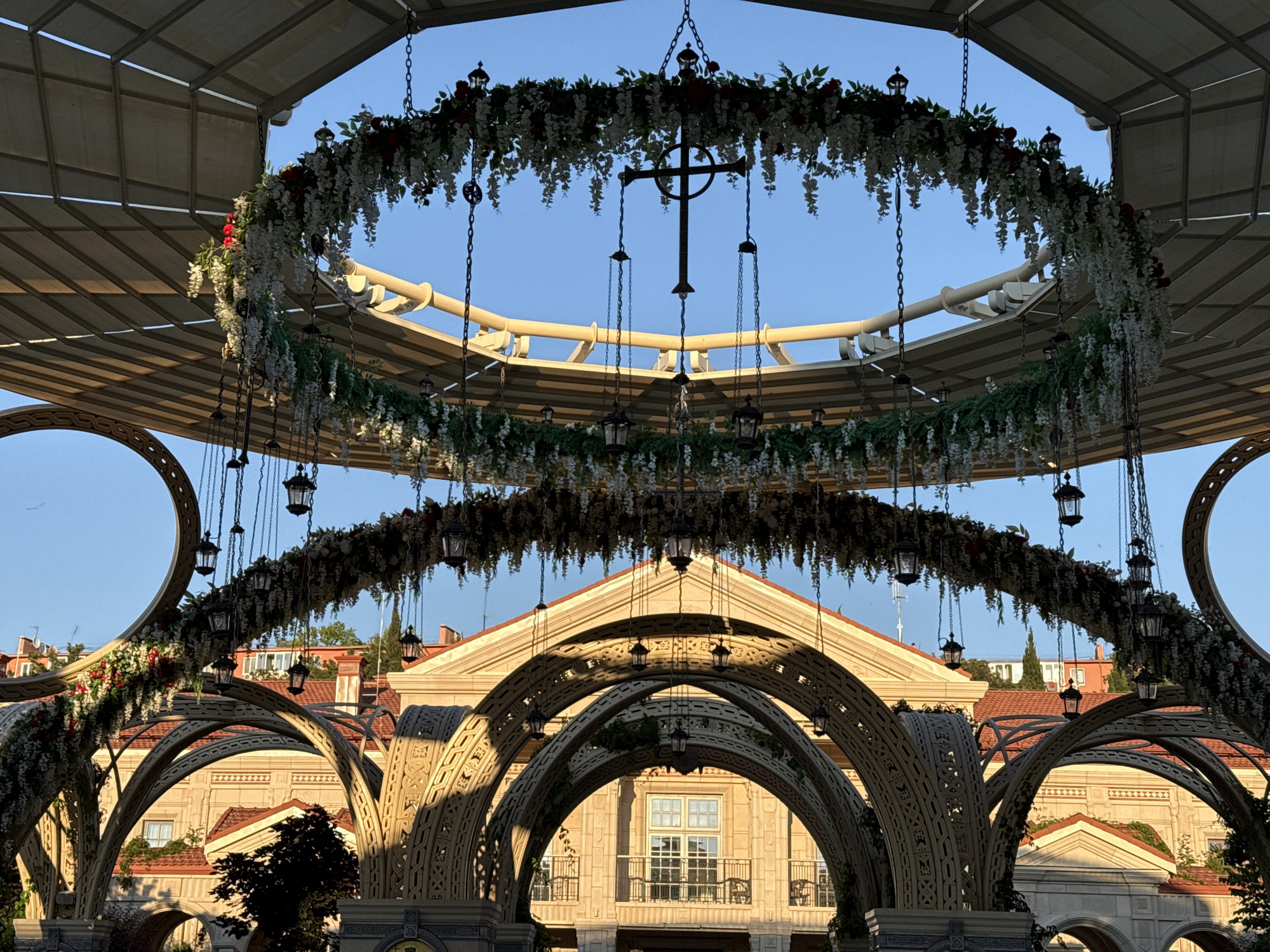 The image shows an outdoor architectural setup with ornate archways and hanging floral decorations. In the foreground, there are intricately designed archways, possibly made of stone or a similar material, forming a passage. These archways are adorned with some greenery and possibly more floral elements.
Above the archways, there are two circular floral arrangements suspended by chains. The upper arrangement features white and red flowers with lush green foliage, and a dark, cross-shaped object hangs from its center, also suspended by chains. Both floral arrangements have small, decorative lanterns hanging from them.
In the background, a building with red-tiled roofs and light-colored walls is visible. The building has multiple windows and balconies. The scene appears to be outdoors, under a light-colored canopy structure, with a clear blue sky in the upper portion of the image. The lighting suggests it might be late afternoon or early morning, with the sun casting shadows and highlighting the architectural details. There are no people visible in the image.