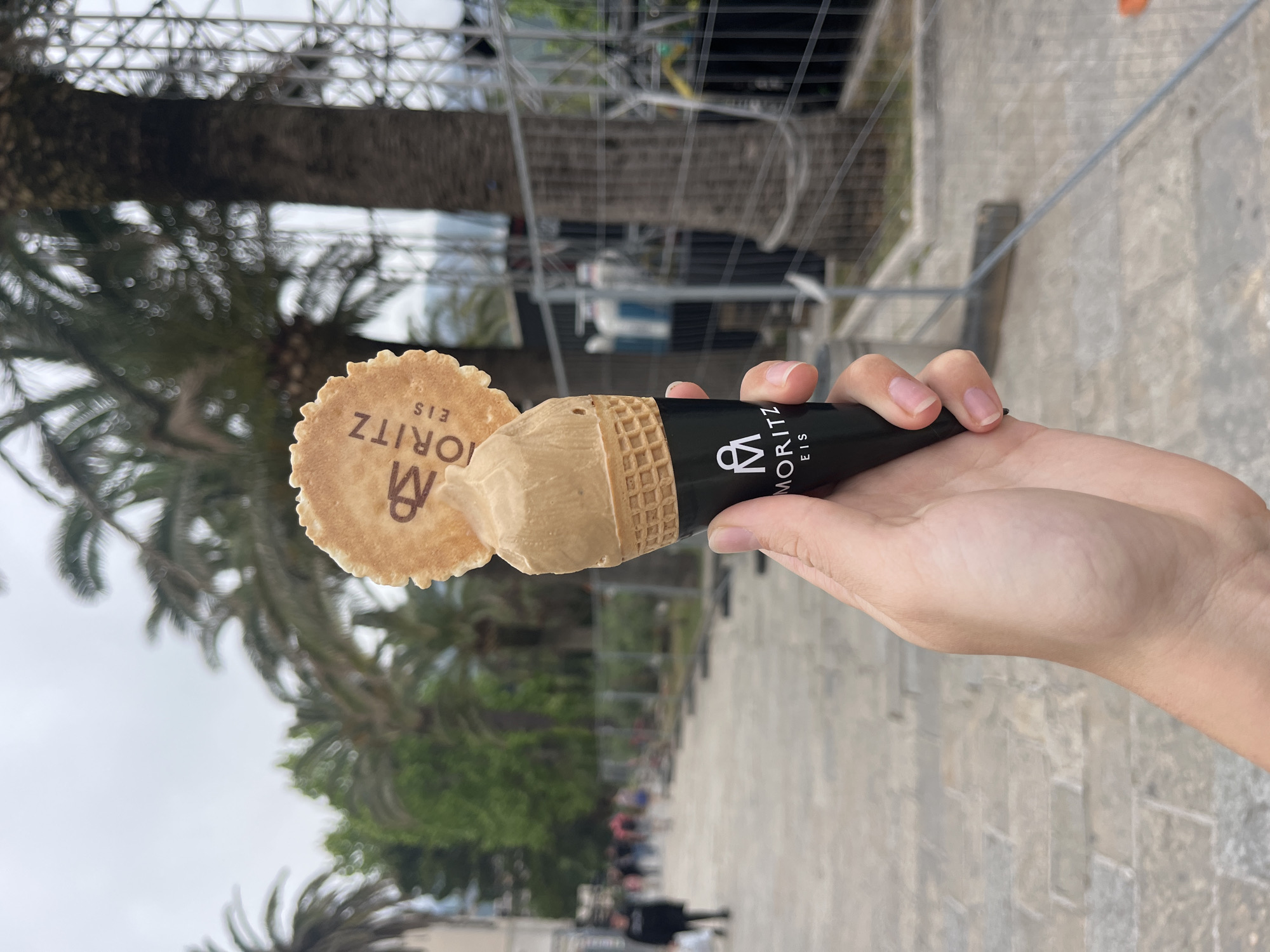 A person holds an ice cream cone with the "Moritz Eis" logo in their right hand. The cone is black with white text, and the ice cream is a light brown color, topped with a round wafer with the same logo. The background includes palm trees and a metal structure. The setting appears to be outdoors on a paved surface, likely within an urban or tourist area. The location is the city of Budva, Montenegro.