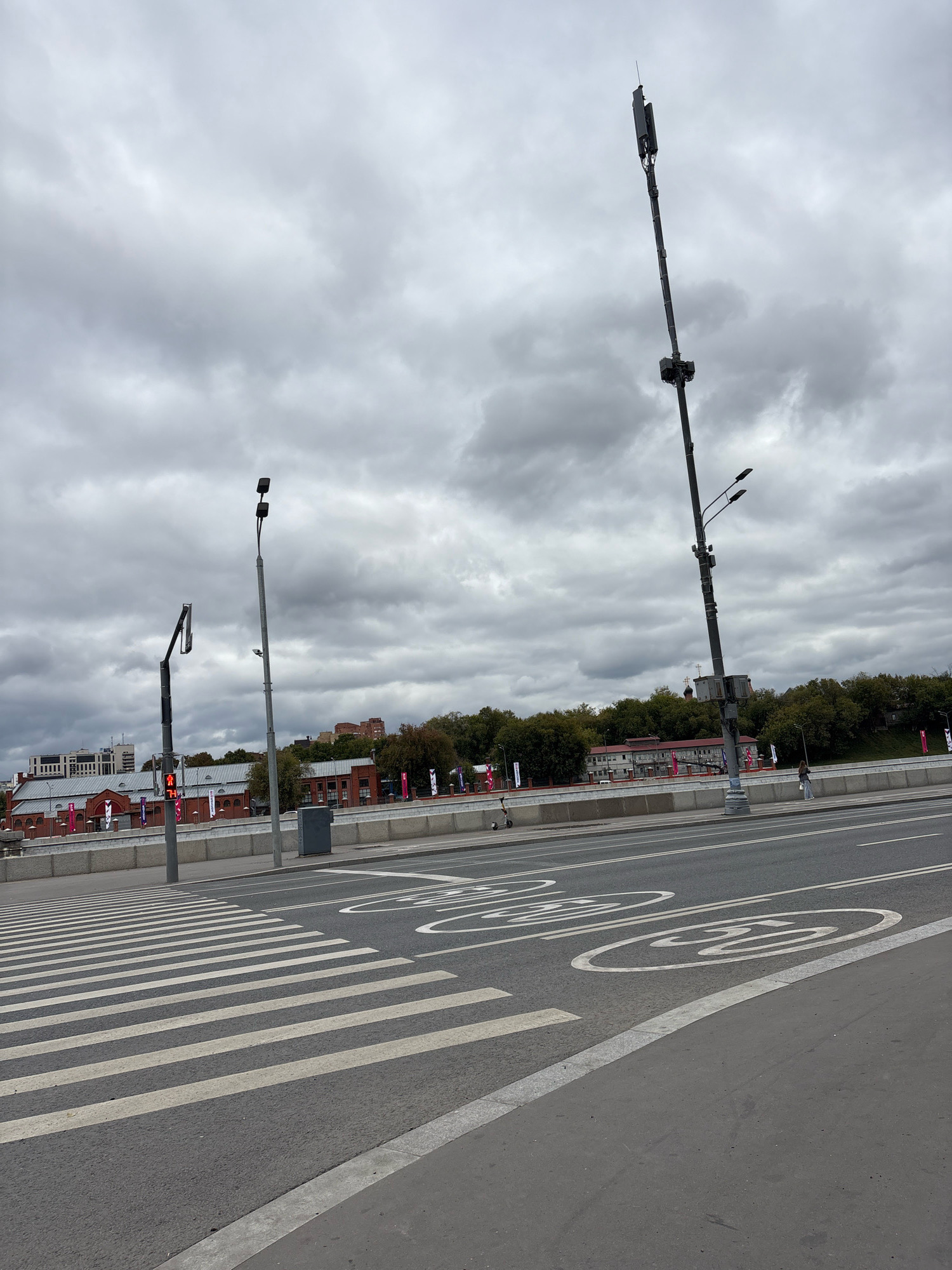 The image shows a street scene on a cloudy day in Moscow, Russia. A wide asphalt road occupies the foreground, with a pedestrian crossing marked by white stripes. To the right of the crossing, speed limit symbols are painted on the road, indicating 30 km/h. A few people and an electric scooter are visible on the sidewalk in the middle ground.
Behind the sidewalk, there is a low concrete barrier, and beyond that, a line of brick buildings with a red roof. Trees with green foliage form a backdrop to the buildings. Several tall communication towers with antennas are present in the scene, one of which is particularly prominent on the right side of the image, extending high into the cloudy sky. Streetlights are also visible. The sky is overcast with heavy grey clouds.