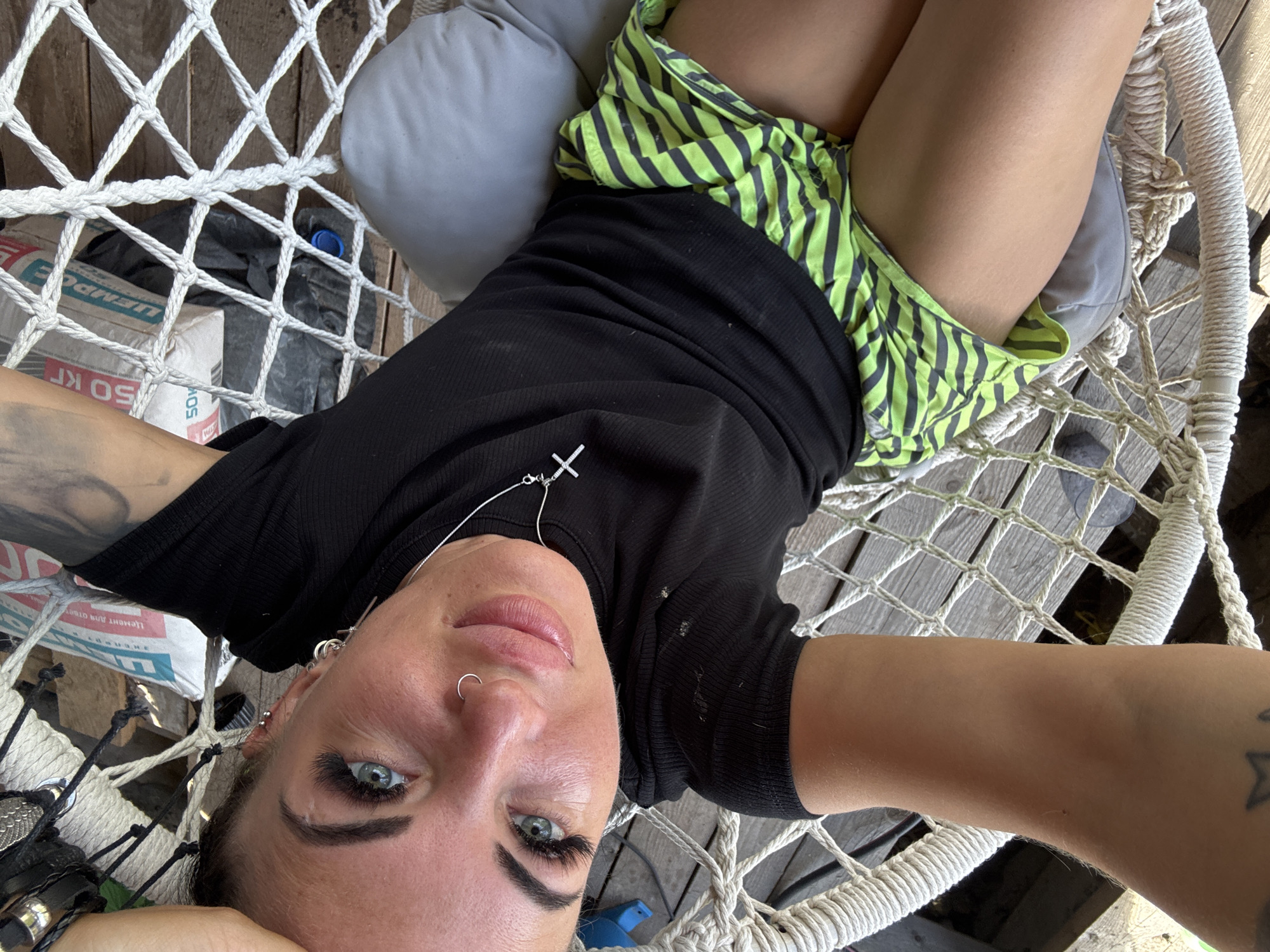 A woman is lying in a hammock, taking a selfie. The woman has long dark hair, blue eyes, and a nose ring. She is wearing a black t-shirt and green and black striped shorts. The hammock is made of white rope and is suspended from wooden beams. There is a gray pillow in the hammock. In the background, there are wooden boards and some bags. The lighting suggests it is daytime and sunny. The woman appears relaxed and happy. Visible text on a bag reads "50 КГ" and "УМЕНЬШЕНО".
