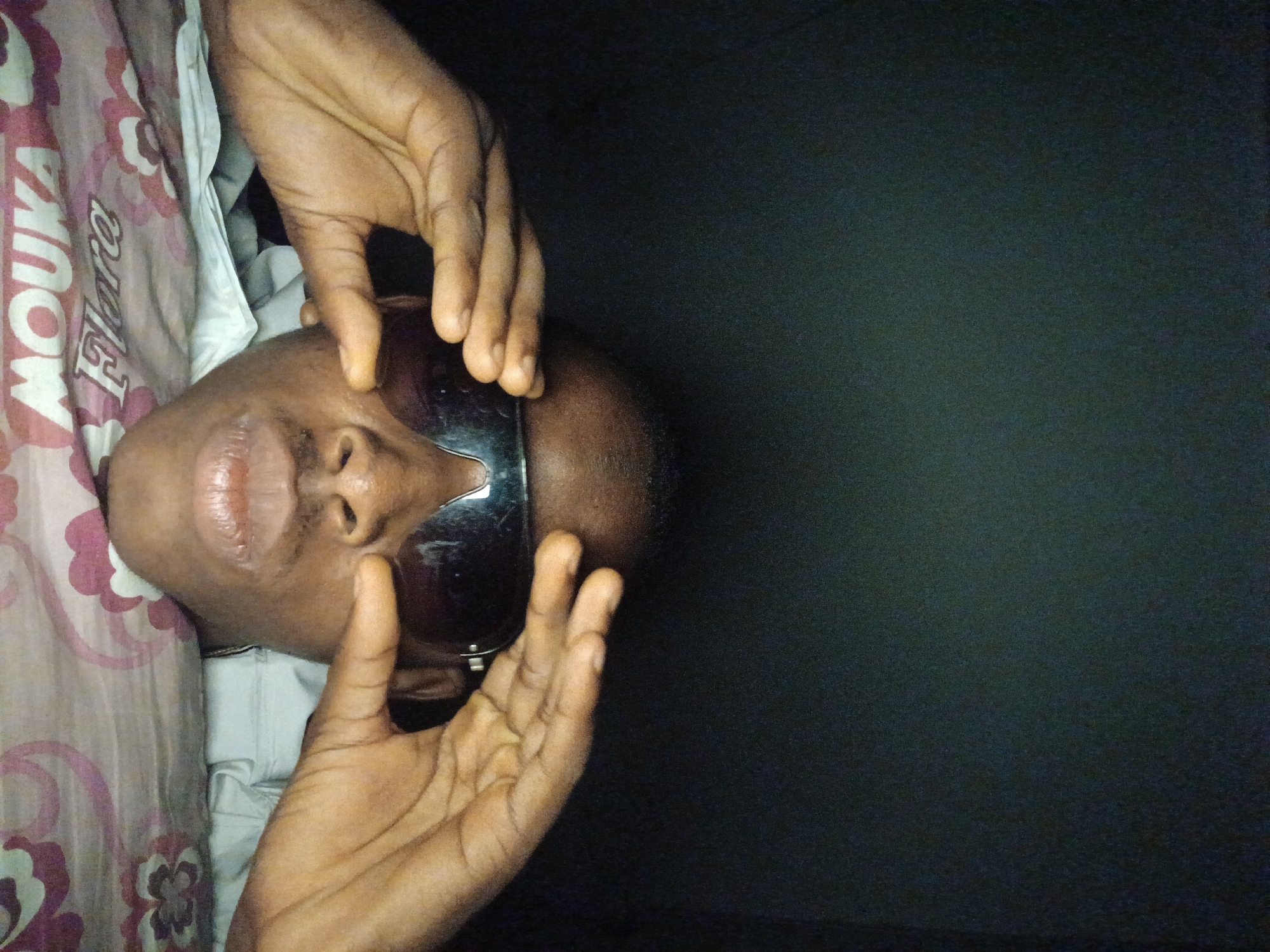 A person wearing sunglasses is lying down, with their hands holding the sunglasses on their face. The background is dark, with a pattern visible on the surface beneath the person. The image is taken from a close-up perspective, focusing on the person's face and hands.