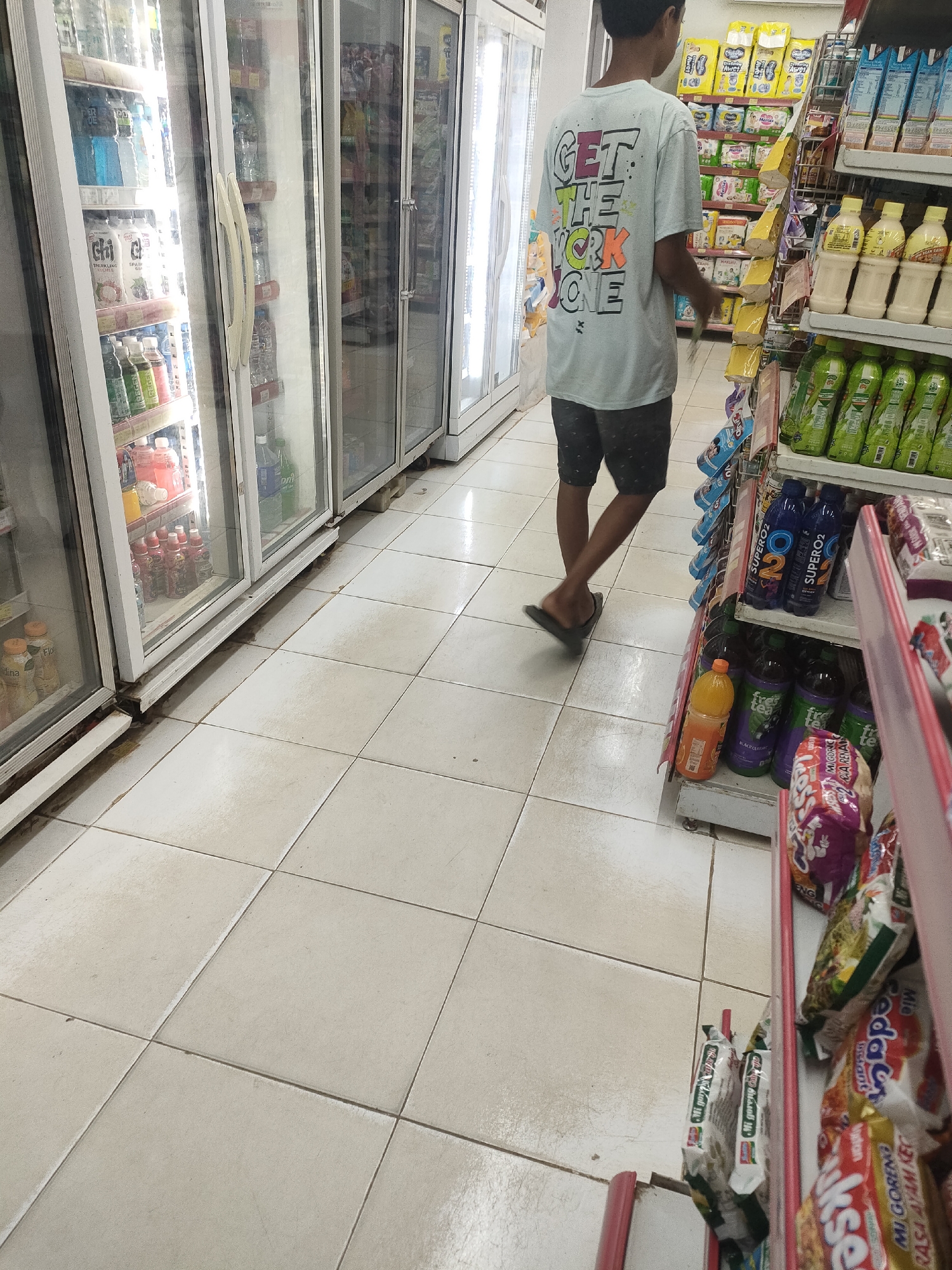 A young person wearing a light-colored t-shirt with a graphic design and dark shorts stands in a convenience store, facing away from the camera. The t-shirt has the text "GET THE WORK DONE" in a colorful, blocky font. The person is wearing dark flip-flops. The store has tiled flooring and rows of refrigerated display cases filled with beverages on the left. On the right, shelves are stocked with various packaged food items and drinks. The overall lighting is bright, typical of a retail environment. The scene appears to be located in Desa Banyumas, Indonesia, as indicated by the products visible on the shelves, which include Indonesian brands such as Indomie.