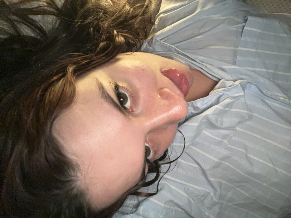 A woman with brown hair is lying on her back, partially on a light blue and white striped shirt. Her head is tilted, and her tongue is extended. Her eyes are open, looking directly at the viewer. Her skin appears moist, and the lighting accentuates the contours of her face. The background is slightly blurred, drawing attention to the woman's face.