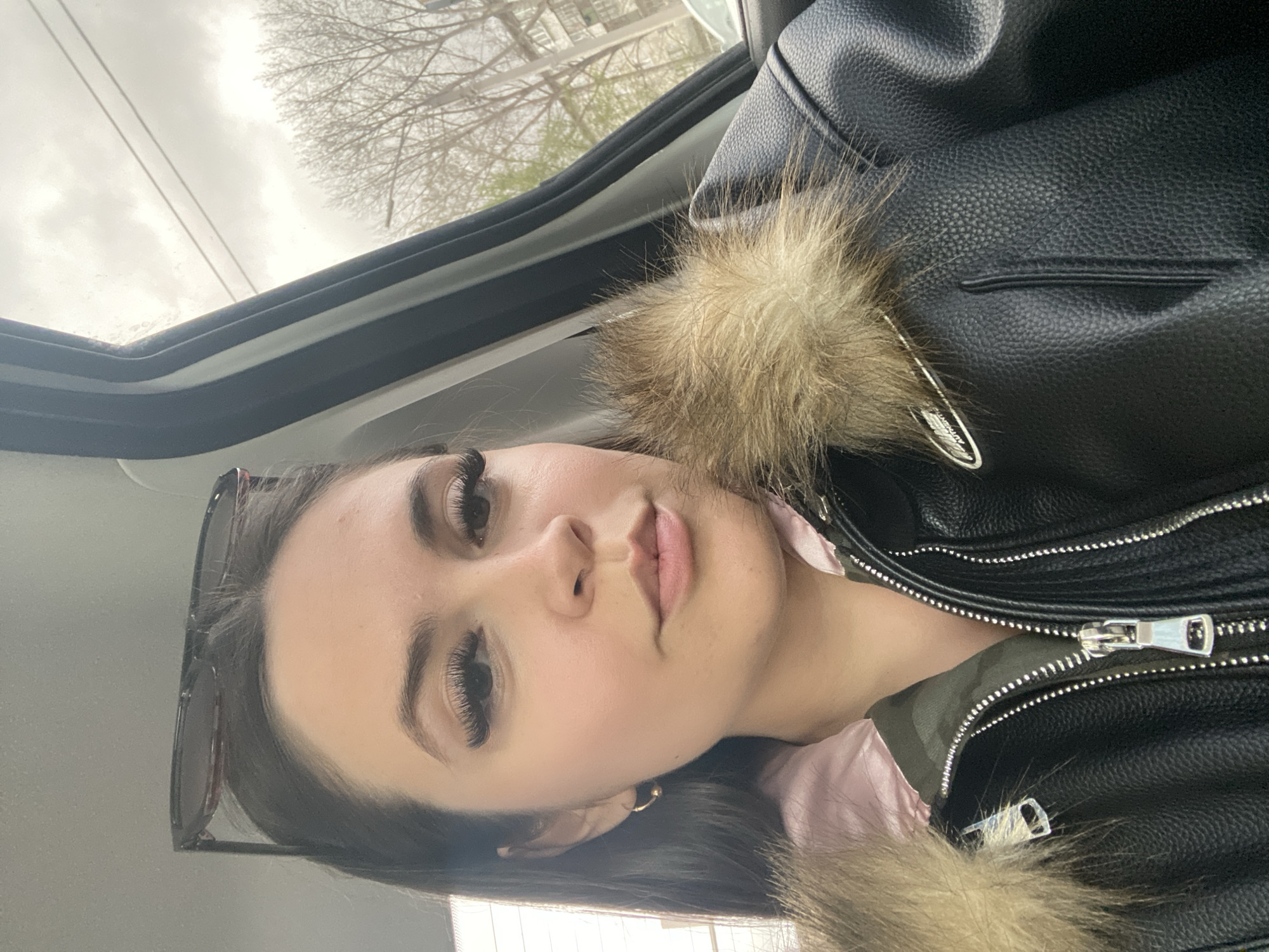 A young woman is in the passenger seat of a car, turned slightly towards the camera. She has dark hair, fair skin, and long eyelashes. Sunglasses are propped on her head. She wears a black leather jacket with a fur collar over a pink shirt. The car's interior is visible, including the roof and window frame. Outside, a cloudy sky and bare tree branches are visible. While the prompt mentions Chaikovsky, Russia, there is no visual information within the image itself that confirms this location.