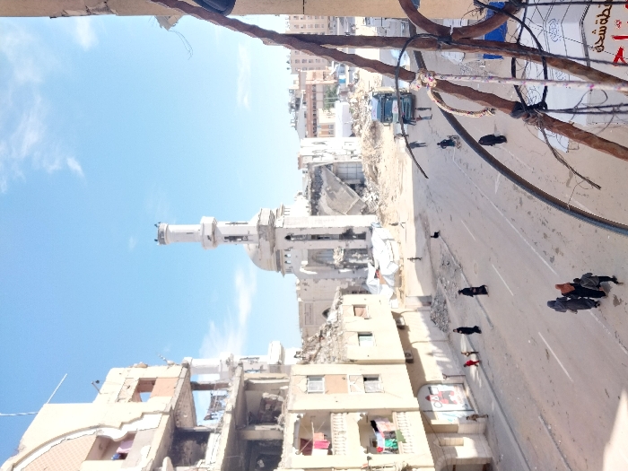 The image displays a wide-angle view of a street in Khan Yunis, Palestinian Territory, characterized by extensive structural damage under a clear blue sky with sparse clouds. Dominating the central background is a severely damaged mosque featuring a white minaret and dome; its main structure shows significant collapse, with interior elements exposed. To the left, a multi-story building, likely residential, exhibits substantial damage, including exposed floors, cracked facades, and laundry items visible on balconies.
The street itself is a wide, unpaved or dirt-covered expanse, showing evidence of vehicular traffic and strewn with debris. Approximately ten to fifteen individuals are present. In the lower mid-ground, a group of three people, including two adults in dark clothing (appearing to wear headscarves) and a child, walk away from the viewer. Other individuals, similarly dressed, are observed walking singly or in small groups along the street and sidewalks. Two children are visible near the damaged building on the left. Further down the street, near the mosque, a large green and white truck is present, with several figures standing near it. In the extreme right foreground, rusty, bent metal rods and electrical wires hang from an unseen structure, partially obscuring a sign with red Arabic script. Piles of rubble and additional damaged buildings are visible in the background, reinforcing the widespread destruction.