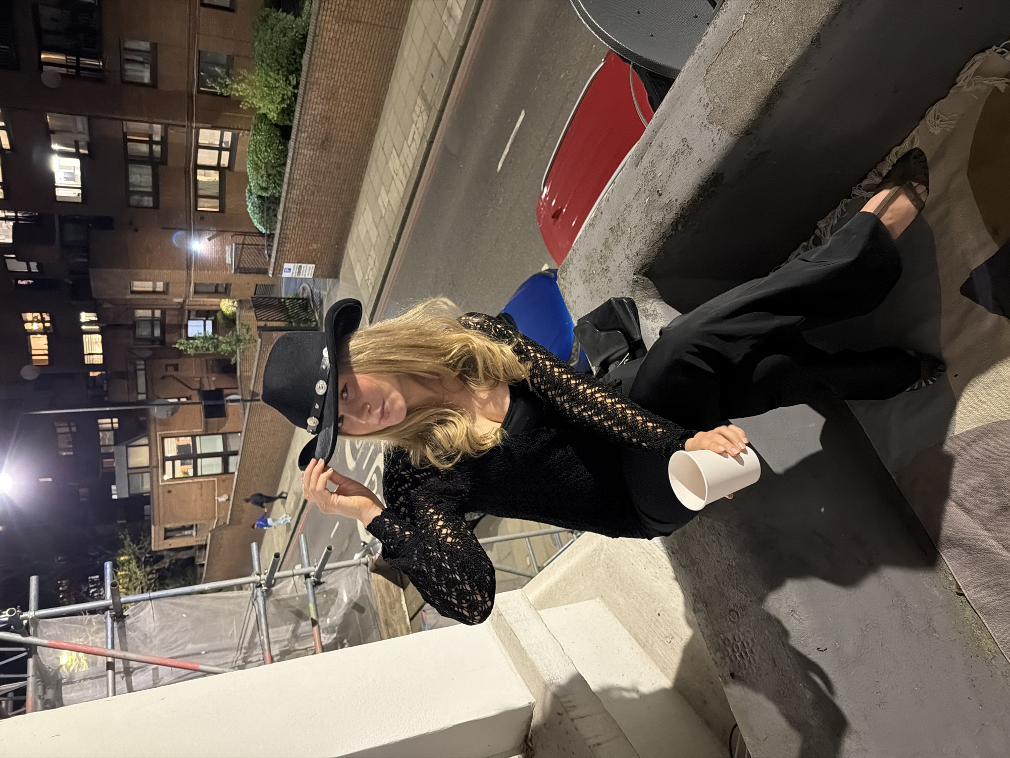 A blonde woman in a black cowboy hat, black lace top, and black pants sits on a concrete ledge at night. She holds a white cup in her right hand. Scaffolding stands in front of a white wall. A street runs behind the ledge with parked cars, a red car, and a blue object. Buildings with lit windows are across the street. The location appears to be a street in London, United Kingdom.