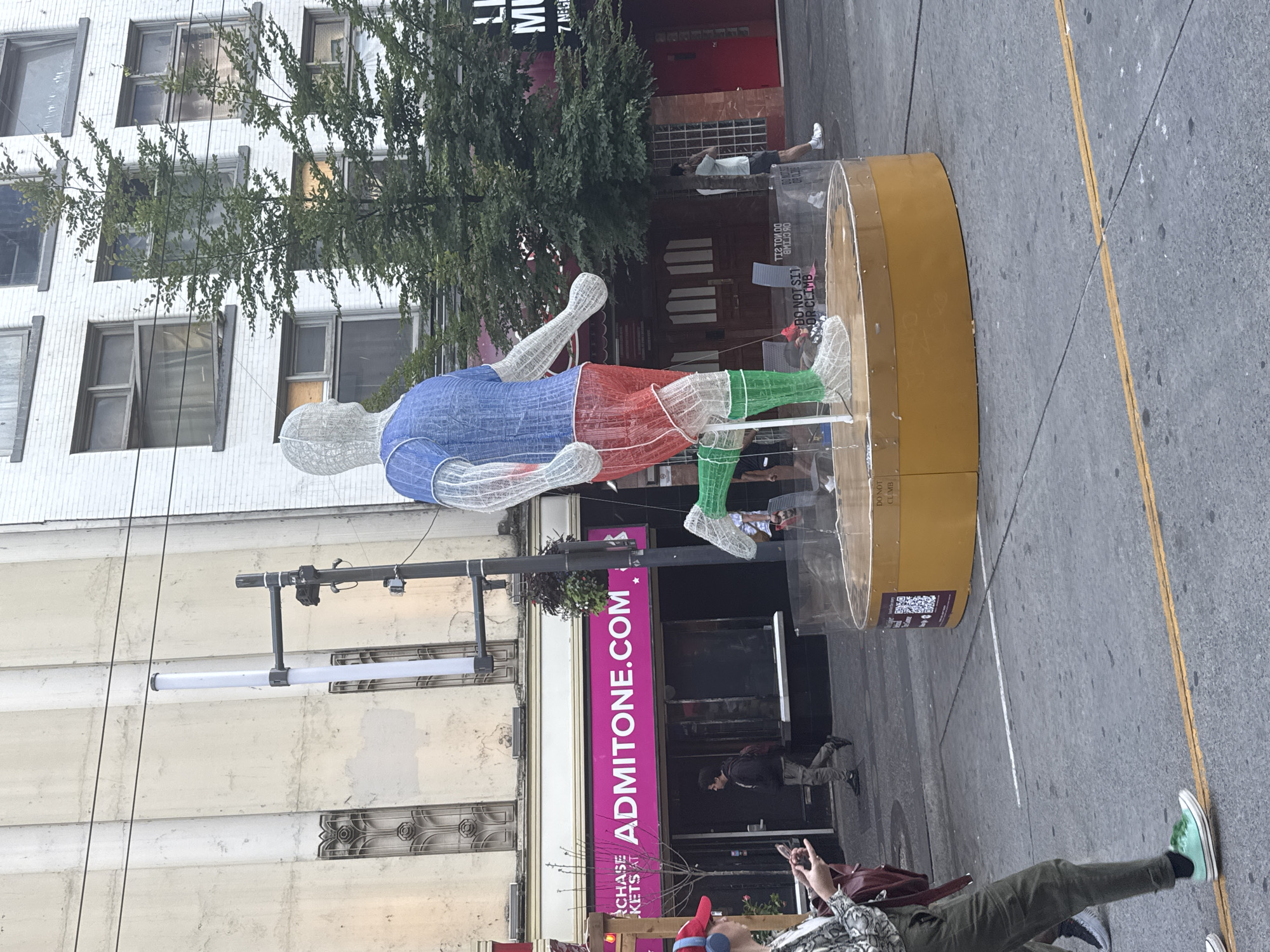 A life-sized sculpture of a runner, constructed from white wire mesh and colored fabric, is displayed on a circular, yellow platform surrounded by a clear plastic barrier. The runner is depicted mid-stride, wearing a blue shirt, red shorts, and green socks. The sculpture is situated outdoors on a street, with a building displaying a large pink sign that reads "ADMITONE.COM" visible behind it. Several people are present in the scene, including a woman in the foreground looking at her phone and wearing a red hat and headphones, and other pedestrians walking in the background. Trees and streetlights are also visible. The overall environment suggests an urban setting, likely a street in Vancouver, Canada, given the style of the architecture and the presence of the "ADMITONE.COM" sign, a known ticketing service in the city.
