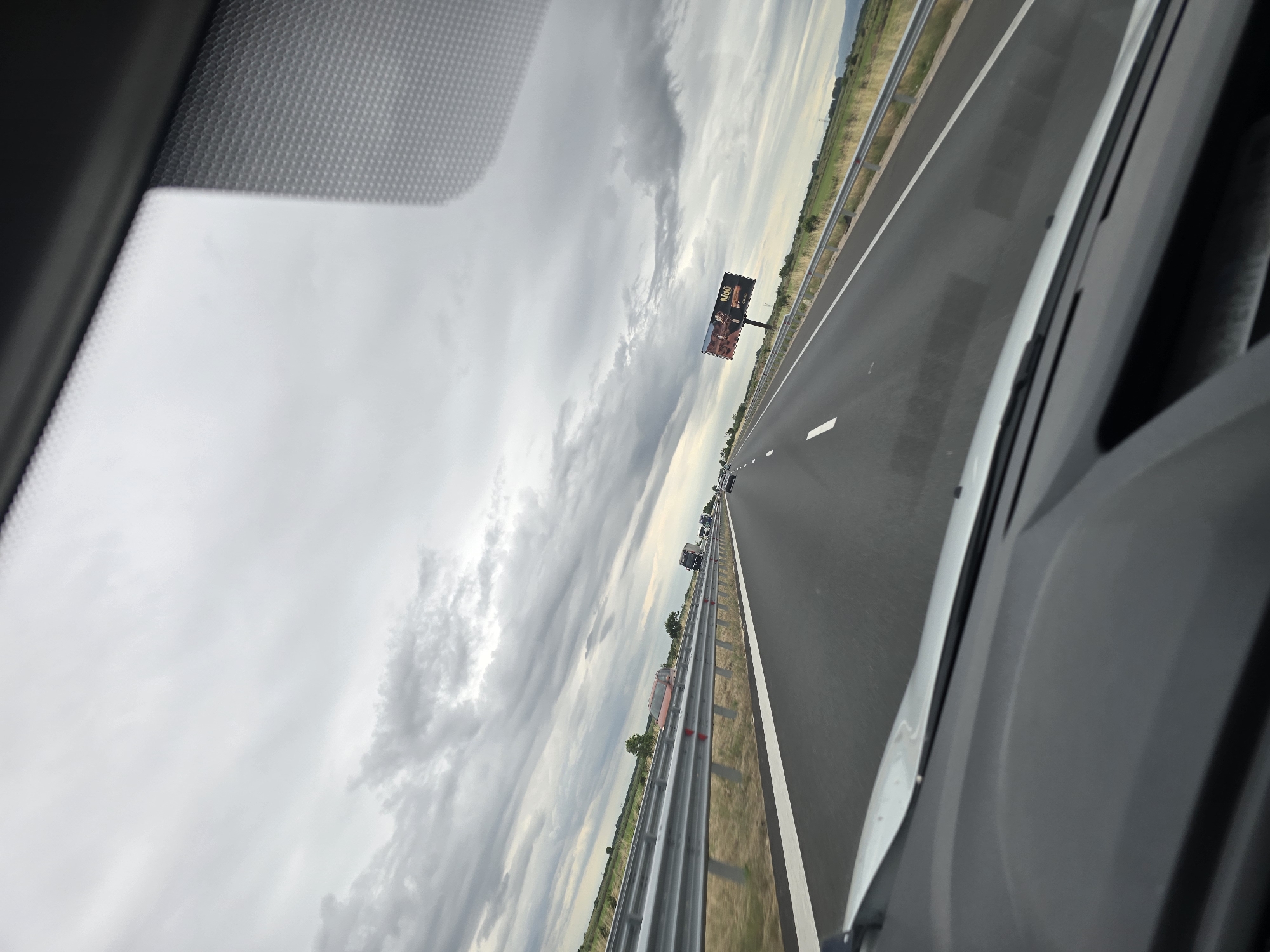 The image captures a view from inside a vehicle traveling on a highway in Malo Konare, Bulgaria. The sky is overcast with heavy, grey clouds. The highway is asphalt with clear white lane markings. On the left side of the road, a metal guardrail separates the highway from a dry, grassy field with sparse vegetation and a few distant trees. On the right side of the highway, there is a similar grassy field with more trees visible in the distance and a billboard advertising what appears to be a product with text that reads "Nuii". Several vehicles, including cars and trucks, are visible on the highway ahead, suggesting moderate traffic. The foreground shows the dashboard and windshield interior of the vehicle.