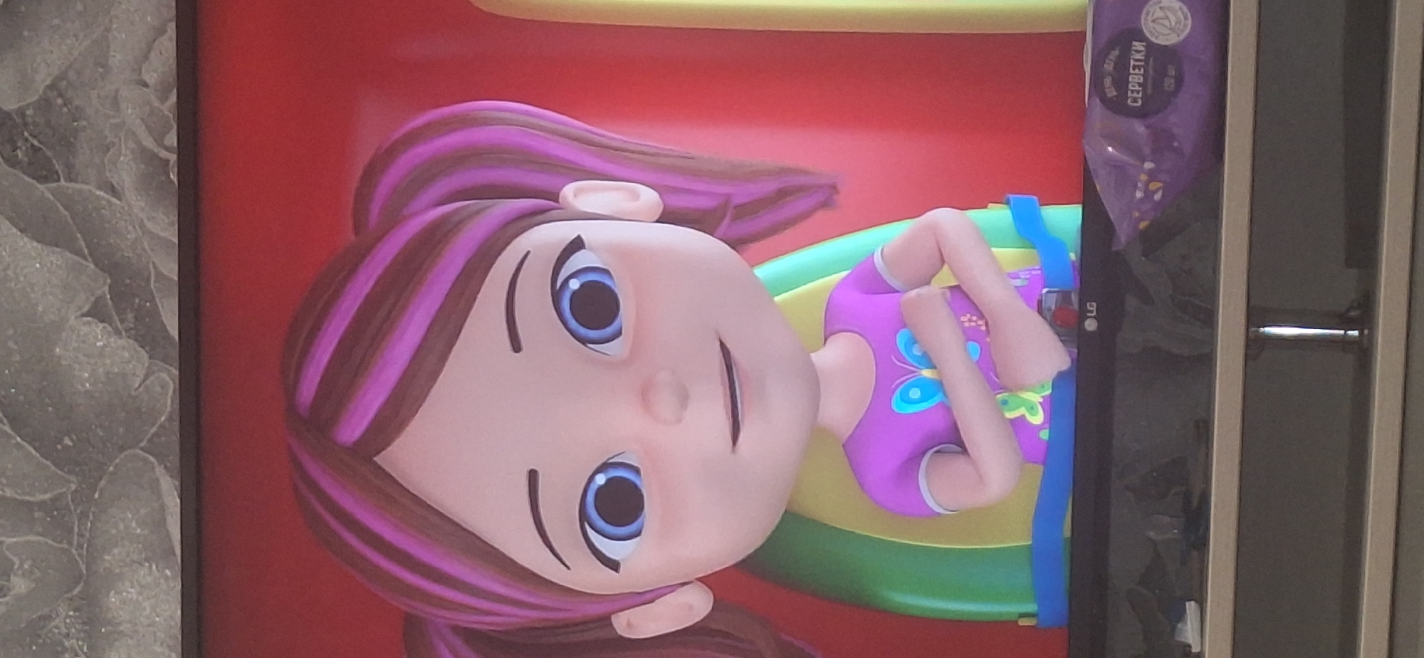 The image shows a close-up of a cartoon character, a young girl with brown hair with purple streaks styled into pigtails. She has large blue eyes and a slight smile. She is wearing a purple t-shirt with a butterfly design. She appears to be sitting in some kind of green and yellow seat with a blue strap across her chest. The background is primarily red. Below the cartoon character, a portion of a television screen is visible, displaying the LG logo. To the right of the LG logo, a purple package of wet wipes is visible with text that appears to be in Cyrillic. The television is sitting on a shelf unit. The wall behind the television has a textured pattern. The photograph appears to have been taken indoors. There is no information in the image to indicate a specific location within Bila Tserkva, Ukraine.