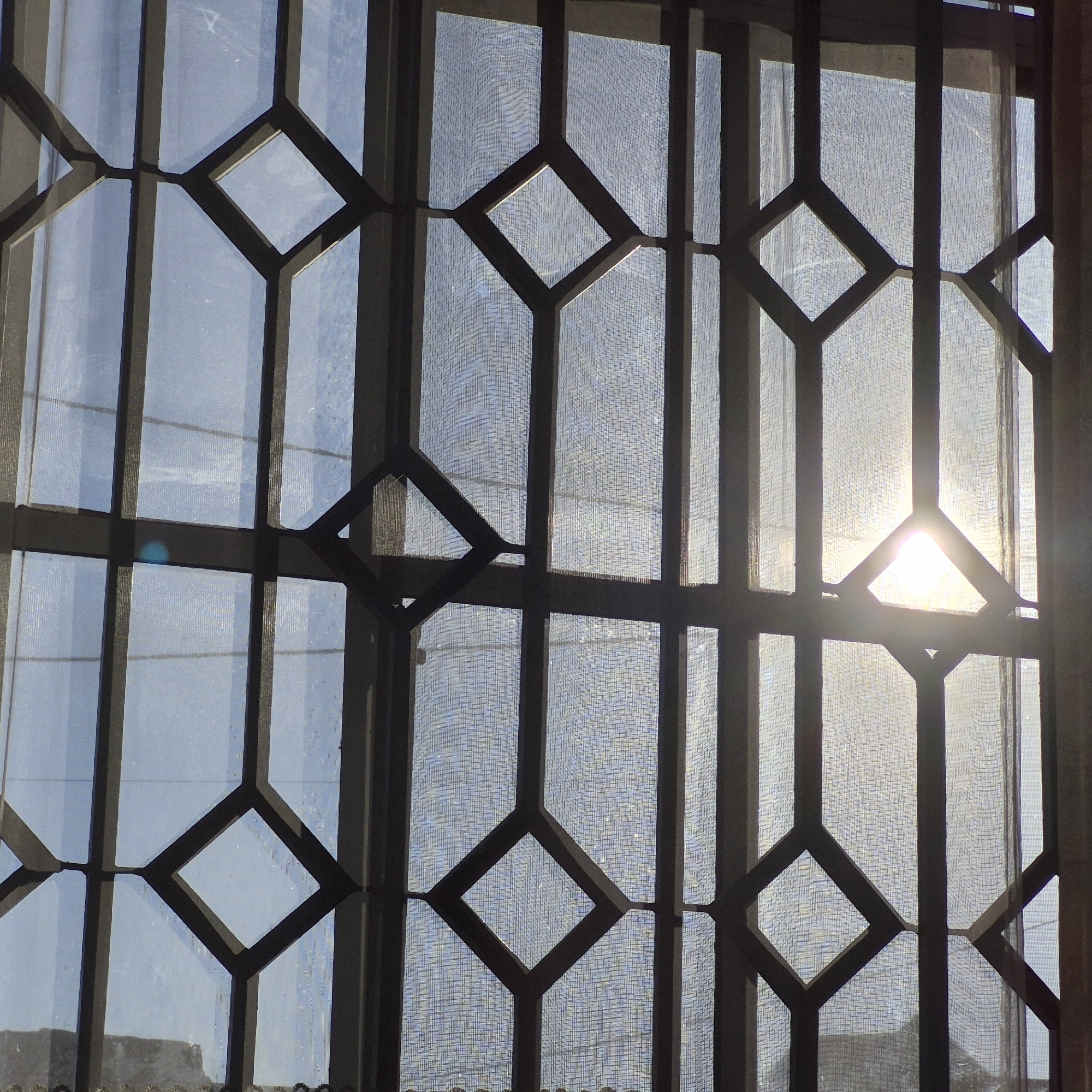 The image shows a window with a decorative metal grate. The grate is composed of vertical bars and horizontal bars that form diamond shapes. A sheer curtain is visible behind the grate, and sunlight is streaming through the window, creating bright highlights. The setting appears to be an interior space looking outwards, possibly a residential or office building. The image captures the geometric patterns of the grate and the interplay of light and shadow. There is no identifiable information in the image to pinpoint its location in Dodoma, Tanzania.