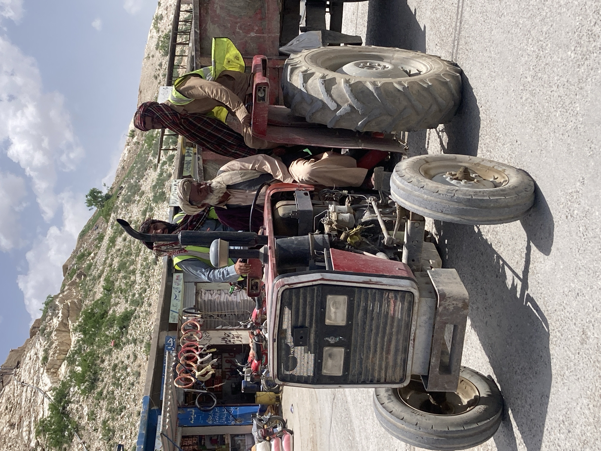 The image captures a bustling roadside scene in Loralai, Pakistan, featuring a prominent red tractor and several individuals. The photograph is taken during bright daylight, evidenced by the clear blue sky with scattered white clouds and the distinct shadows cast on the paved ground.
In the foreground, a robust red tractor is parked. An older man with a long white beard and a white turban is seated on the tractor's engine area, wearing a light-colored tunic. Behind him, another man, sporting a red and plaid head covering, is positioned on the rear fender, clad in a brown shirt and a bright yellow safety vest. A third man, also wearing a yellow safety vest over a blue shirt, is visible near the front left side of the tractor, holding onto a pipe or handle. The tractor has large, heavily-treaded rear tires and smaller front tires, with its distinctive front grille and headlights clearly visible.
To the left of the tractor, a series of small, informal roadside shops or stalls are lined up. These shops display various items, including what appear to be hanging bicycle parts or household goods. Signs on these shops feature text in an unidentifiable script, likely Urdu or Pashto.
The background is dominated by a rugged, arid mountain or hillside, characterized by rocky surfaces and sparse patches of green vegetation. The scene conveys an everyday moment in a rural or semi-urban setting, with people gathered around a functional vehicle against a natural, mountainous landscape. Emotions are not distinctly discernible, but the scene appears calm.