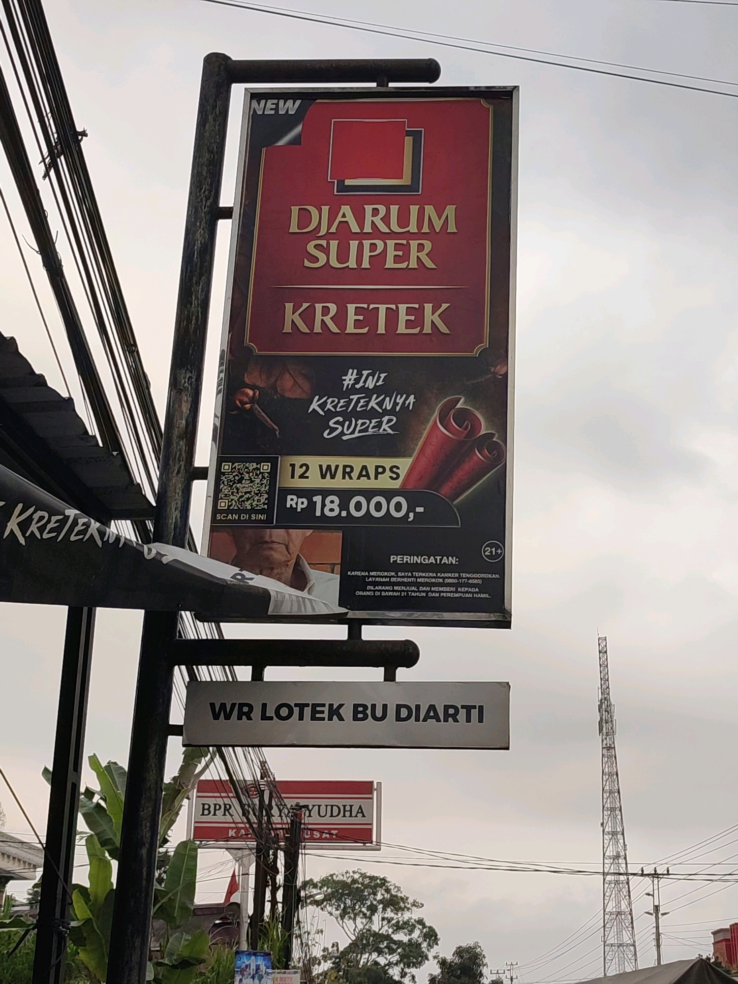 The image shows a vertical advertisement for "DJARUM SUPER KRETEK" cigarettes, featuring the text "NEW" in the upper left corner and a warning that it is for 21+ individuals. The ad prominently displays the brand name, "DJARUM SUPER KRETEK," the product's count "12 WRAPS," and its price "Rp 18.000,-." There is a QR code for scanning. Below this advertisement, another sign reads "WR LOTEK BU DIARTI." Further down, a red and white sign for "BPR KARYUDHA" is visible. The scene is outdoors, with power lines running across the frame and a cell tower in the background. The sky is overcast. The location is identified as Karangluhur, Indonesia.