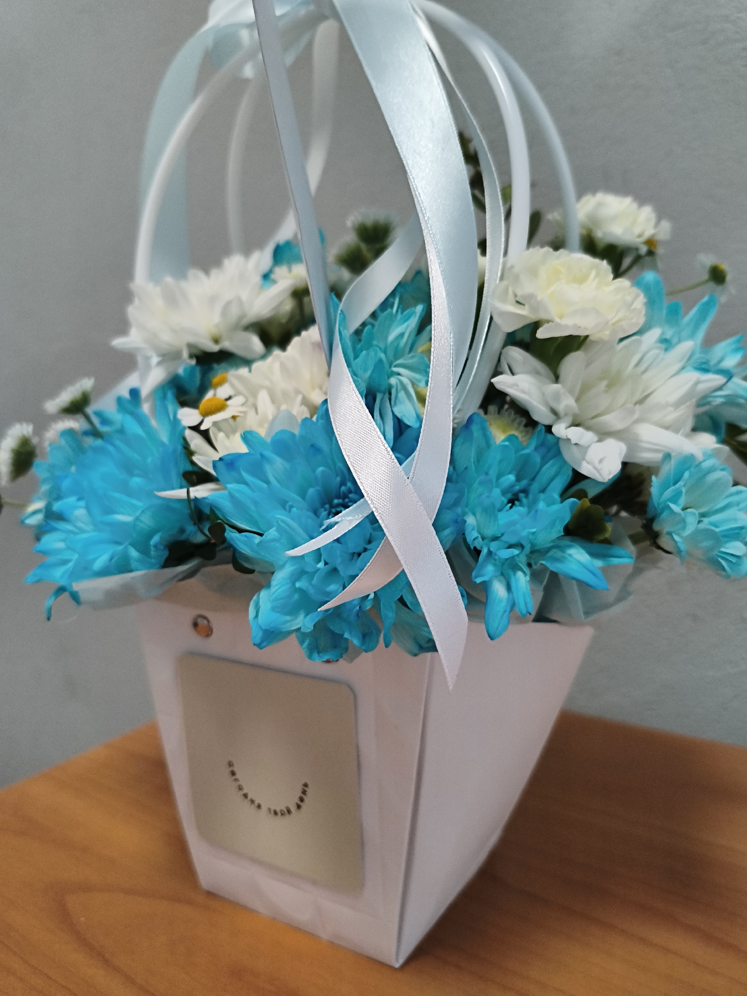 The image shows a close-up of a white paper flower arrangement box filled with blue and white chrysanthemums. The box has a metallic plaque on the front with text that is difficult to read but appears to be a smiley face. The box is adorned with light blue and white ribbons. The arrangement is placed on a wooden surface, likely a table, with a gray wall in the background. There are no people or animals present in the image. The location is identified as Novaya Usman, Russia.