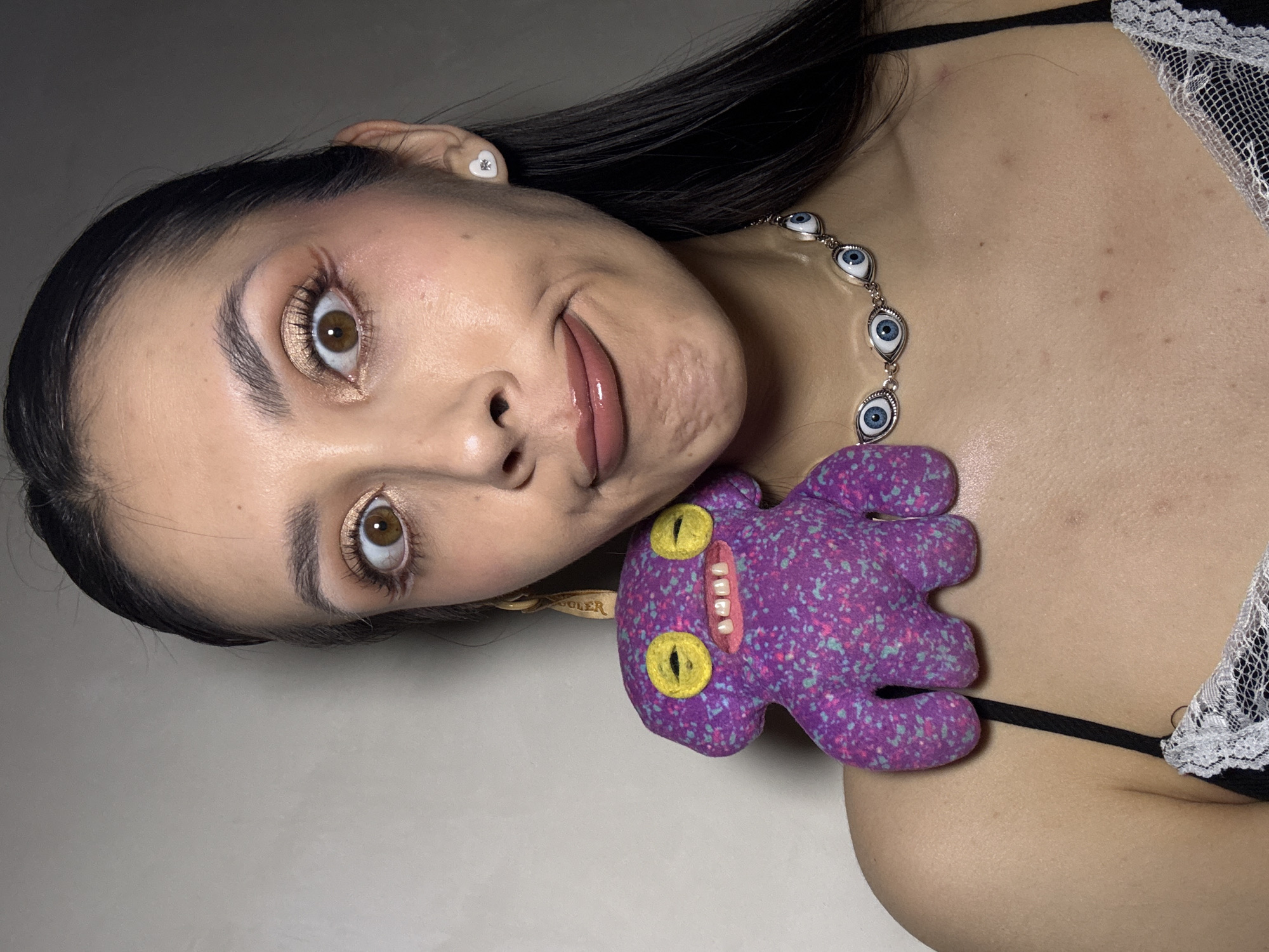 A woman is lying down, facing the camera. She has dark hair, light skin, and is wearing makeup including gold eyeshadow and lip gloss. She is wearing a black top with white lace trim. She has a necklace with eye-shaped beads and a plush, purple, monster-shaped pendant with yellow eyes and a white-toothed grin. She wears a small, heart-shaped earring in her left ear. The background is a plain, light grey surface. There is no indication of the location being Votkinsk, Russia.