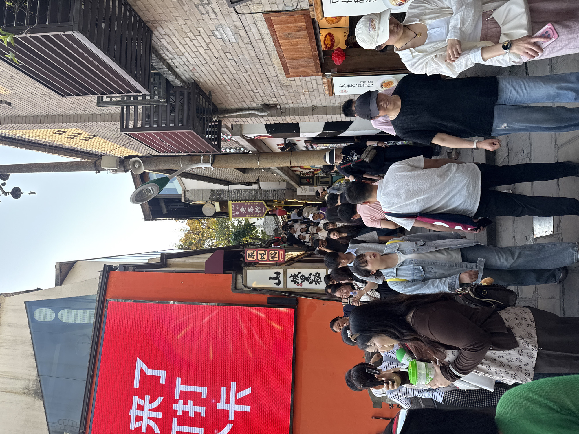 A large group of people are walking along a narrow street in Yuzhong District, China. The people are of Asian descent, dressed in casual clothing, and appear to be tourists visiting the area. There are buildings and shop signs visible along the street. The photograph includes the display for the TEA NING TOWN shop, the 'Lao Cha Guan' tea house, and the shop for 'Yuanshan Chengli Mi' sweets. There are red banners, a street lamp, and other signage in the image.