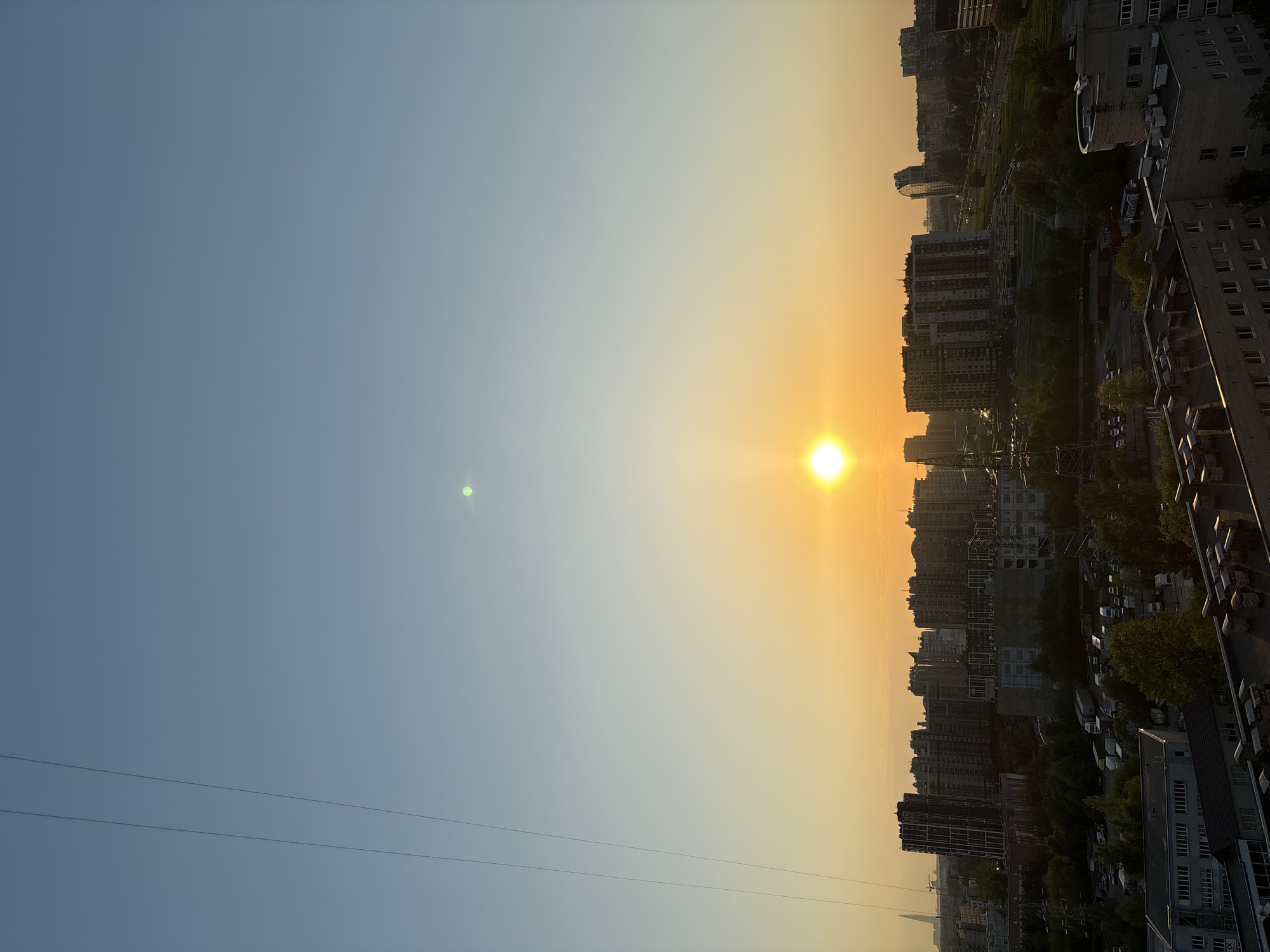 Here is a description of the image:
The image presents a cityscape at sunset. The sky transitions from a light blue to a golden yellow near the horizon where the sun is setting. Buildings line the horizon, some appearing as silhouettes against the bright light of the sun. The buildings vary in height, creating an uneven skyline. The angle of the photograph suggests it was taken from a high vantage point, possibly from the roof of another building. Power lines are visible in the sky. The environment suggests an urban setting.