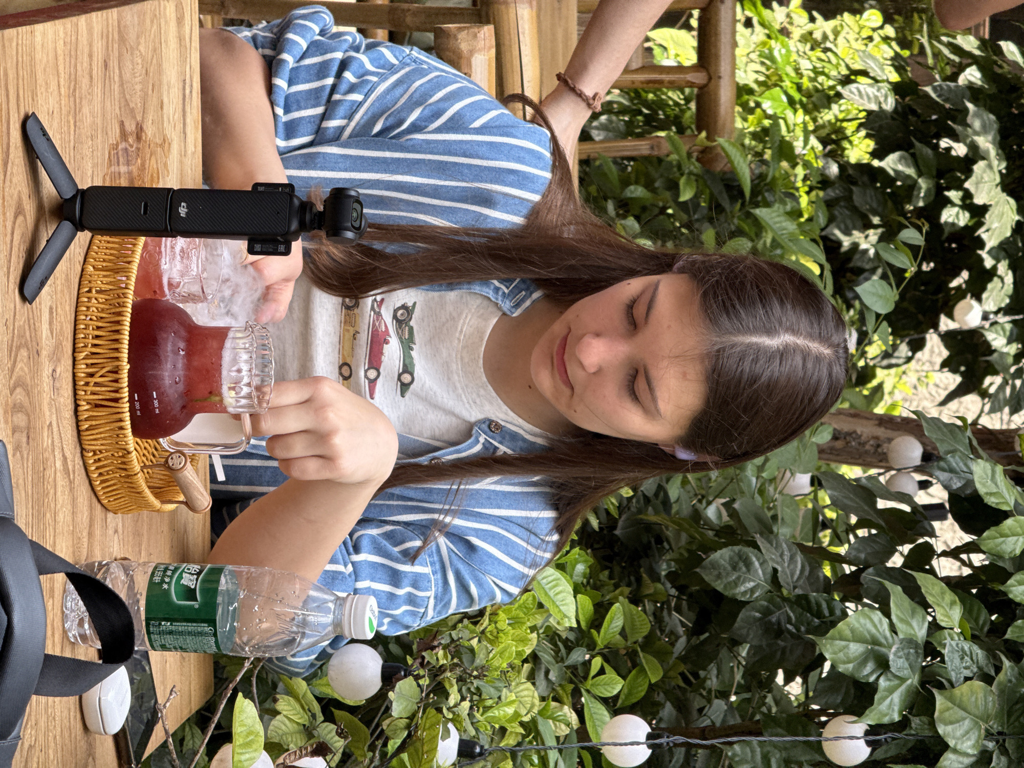 An image, captured in what appears to be Yuzhong District, China, shows a young person with long brown hair, seated at a wooden table in an outdoor or semi-outdoor setting. She wears a light-colored t-shirt featuring a graphic print of cars and a blue and white striped shirt or jacket over it. Her head is tilted downwards, and her eyes are closed or intently focused as she holds a glass mug, seemingly preparing to pour or having just poured a red, iced beverage from a glass pitcher.
On the table, a black DJI Osmo Pocket-like camera device, marked with the "DJI" logo and "EAC," is attached to a small tripod. A woven basket sits near the pitcher. A plastic water bottle with a green label, displaying Chinese characters including "饮用天然水" (Drinking Natural Water) and "500ml," along with the "GongFu" brand, is also on the table beside a black bag strap. The glass pitcher has measurement markings for "200 ml" and "300 ml."
The background is filled with abundant green foliage, suggesting a garden or patio ambiance. White spherical string lights are visible among the leaves and also on the table, contributing to a decorative atmosphere. The natural, bright lighting indicates it is daytime, capturing a tranquil and casual moment.