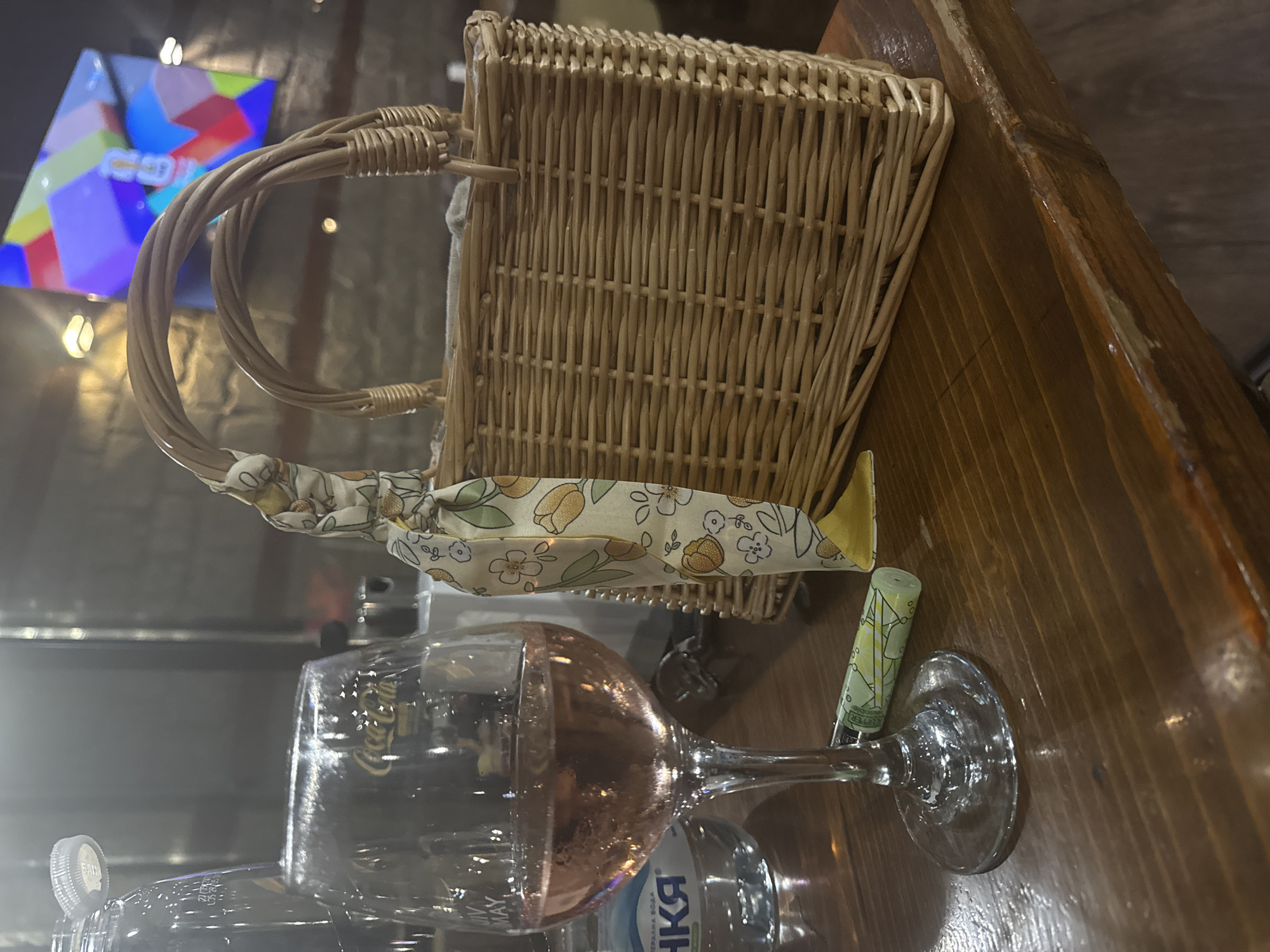 The image depicts a close-up view of a wooden table, likely in an establishment in Rakovski, Bulgaria. In the foreground, a wine glass filled with rose-colored liquid is prominent. Beside it, a small, decorative basket with woven handles and a light-colored fabric adorned with floral patterns rests on the table. A green cylindrical object, possibly a lighter, lies on the table near the base of the wine glass. In the background, a bottle of water with a visible label is partially obscured. Further in the background, a television screen displays a colorful abstract pattern. The overall setting appears to be a dimly lit indoor space, suggesting an evening or nighttime scene.