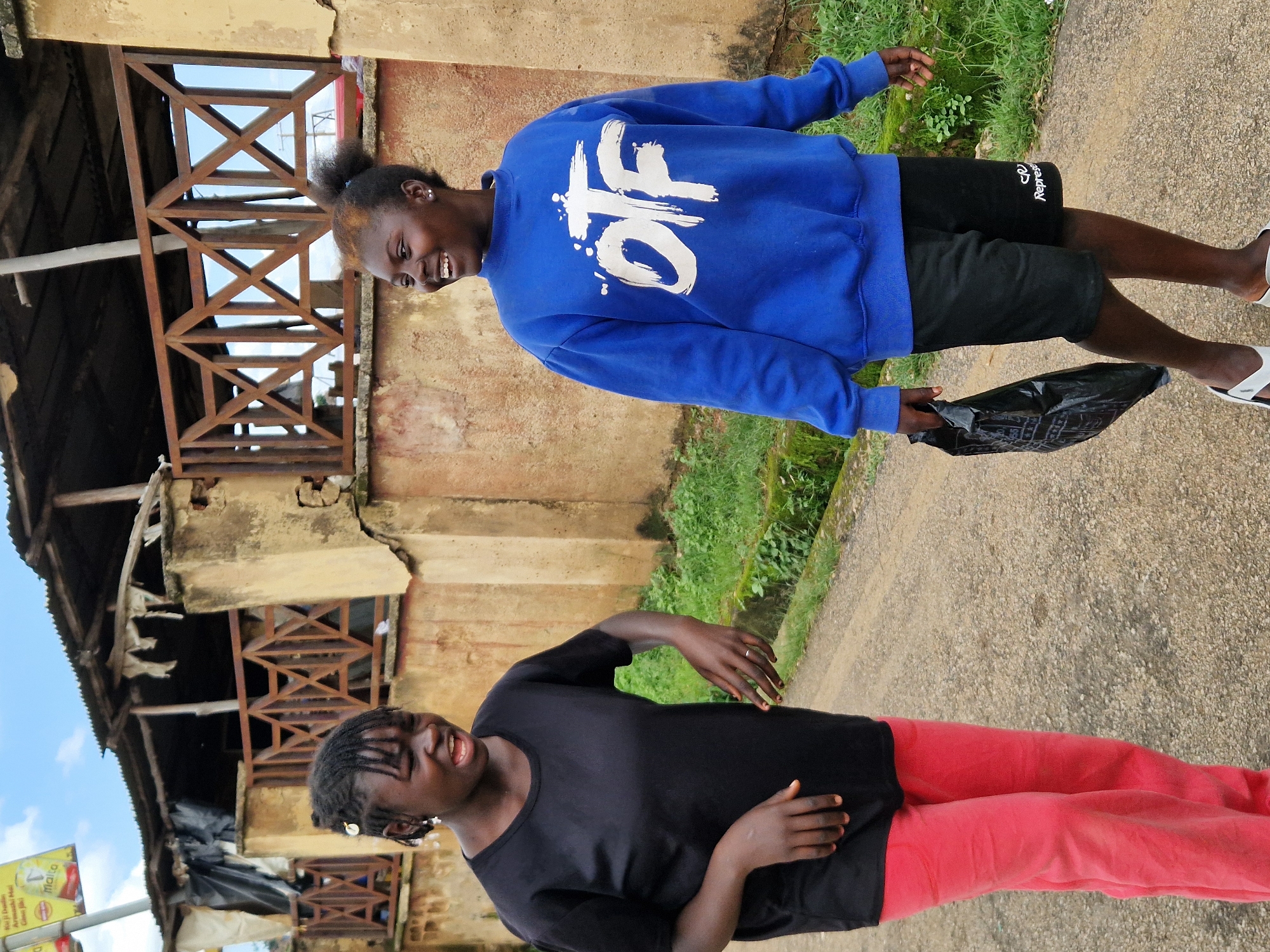 Two young Black females are walking on a dirt road outdoors. The female on the left is wearing a black t-shirt and pink pants. She has her mouth open as if speaking and her left hand is extended. The female on the right is wearing a blue sweatshirt with white lettering and black shorts. She is smiling and looking to her left. She is carrying a black plastic bag in her right hand. In the background, there is a dilapidated building with a corrugated metal roof and wooden railings. The area is overgrown with green grass. The sky is partly cloudy.