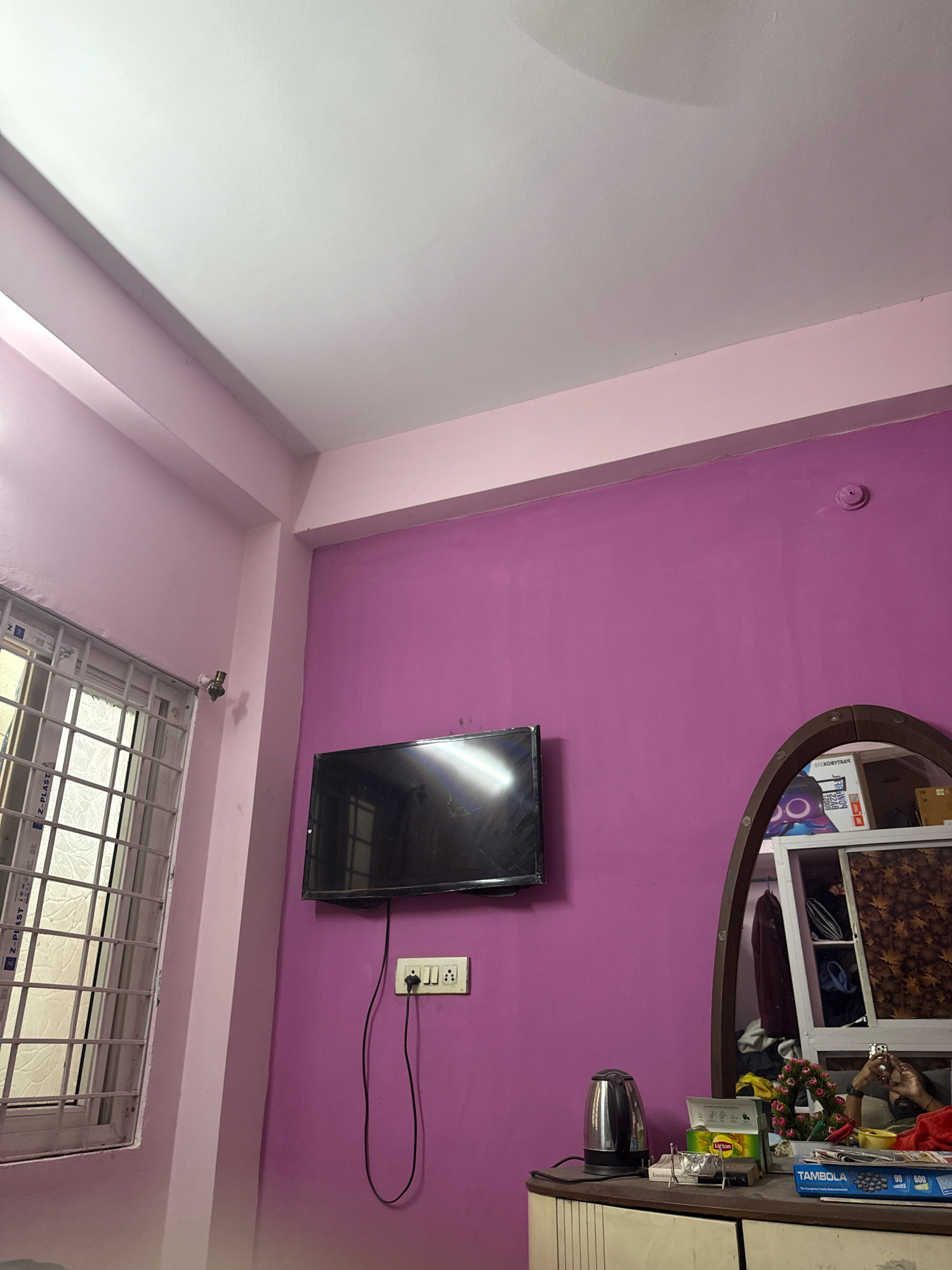 The image depicts an indoor scene, likely a bedroom, with prominent purple walls and a lighter pink accent near the ceiling. On the wall, a flat-screen television is mounted. Below the television, a white electrical outlet with a black cord is visible. To the left of the television, a window with a white metal grille is present. In the foreground, a dresser or cabinet is partially visible, with various items on top. These include a silver electric kettle, a box of Lipton tea, and a box labeled "TAMBOLA." A mirror, oval-shaped with a brown frame, is also present, reflecting items within a closet or shelf. The ceiling is white and appears to have some minor imperfections. The overall impression is of a functional, lived-in space. The specific location in Hyderabad, India, cannot be determined from the visual information provided in the image.