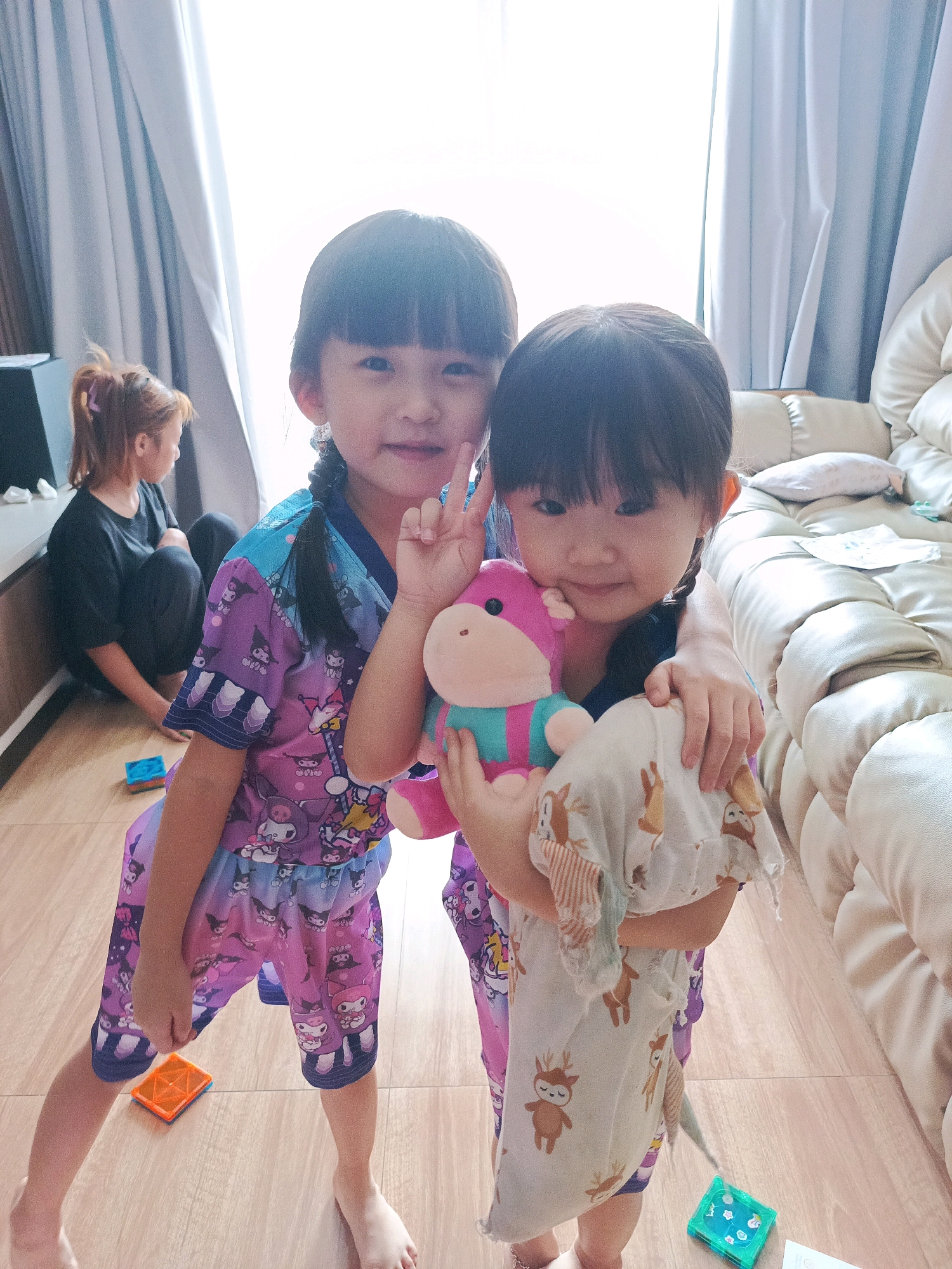 Two young girls are standing in a room in Curug Sangereng, Indonesia. The girl on the left is wearing a purple pajama set with cartoon characters on it. She has black hair in pigtails and is holding a pink and blue stuffed animal. The girl on the right is also wearing pajamas with cartoon characters and is holding a light-colored blanket with deer prints. In the background, another person is sitting on the floor near a cabinet. The room appears to be a living area, with a large beige sofa visible on the right. The floor is made of wood.