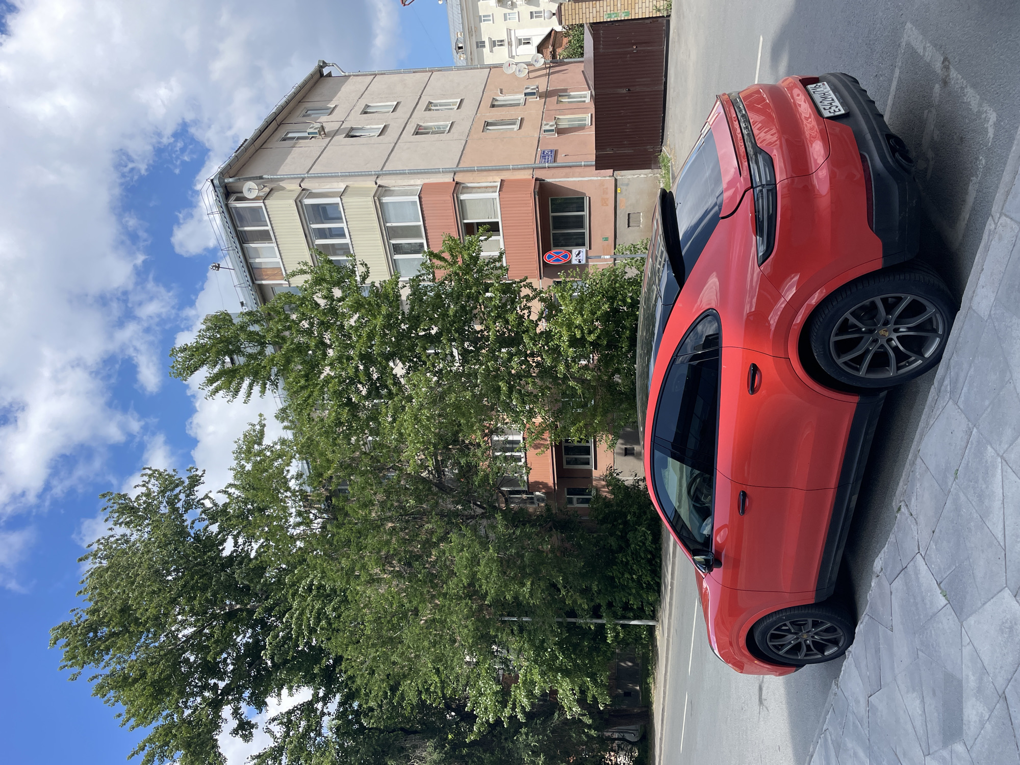 A red Porsche Cayenne Coupe is parked on the side of a street in Kazan, Russia. The car is viewed from its rear passenger side, with the front of the vehicle facing to the left. It is a sunny day, and the sky is blue with scattered white clouds.
The car is parked along a sidewalk paved with grey hexagonal tiles. A paved road with white lane markings is to the left of the sidewalk. Behind the car, a multi-story apartment building with a light beige facade and peach-colored balconies stands. Trees with lush green foliage are in front of the building.
A brown metal gate and a brick fence are visible to the right of the apartment building. A street sign with the number "21" is affixed to the building. A red and white traffic sign depicting a car being towed is also present. The license plate of the Porsche reads "E542MH 716". There are no people visible in the image.