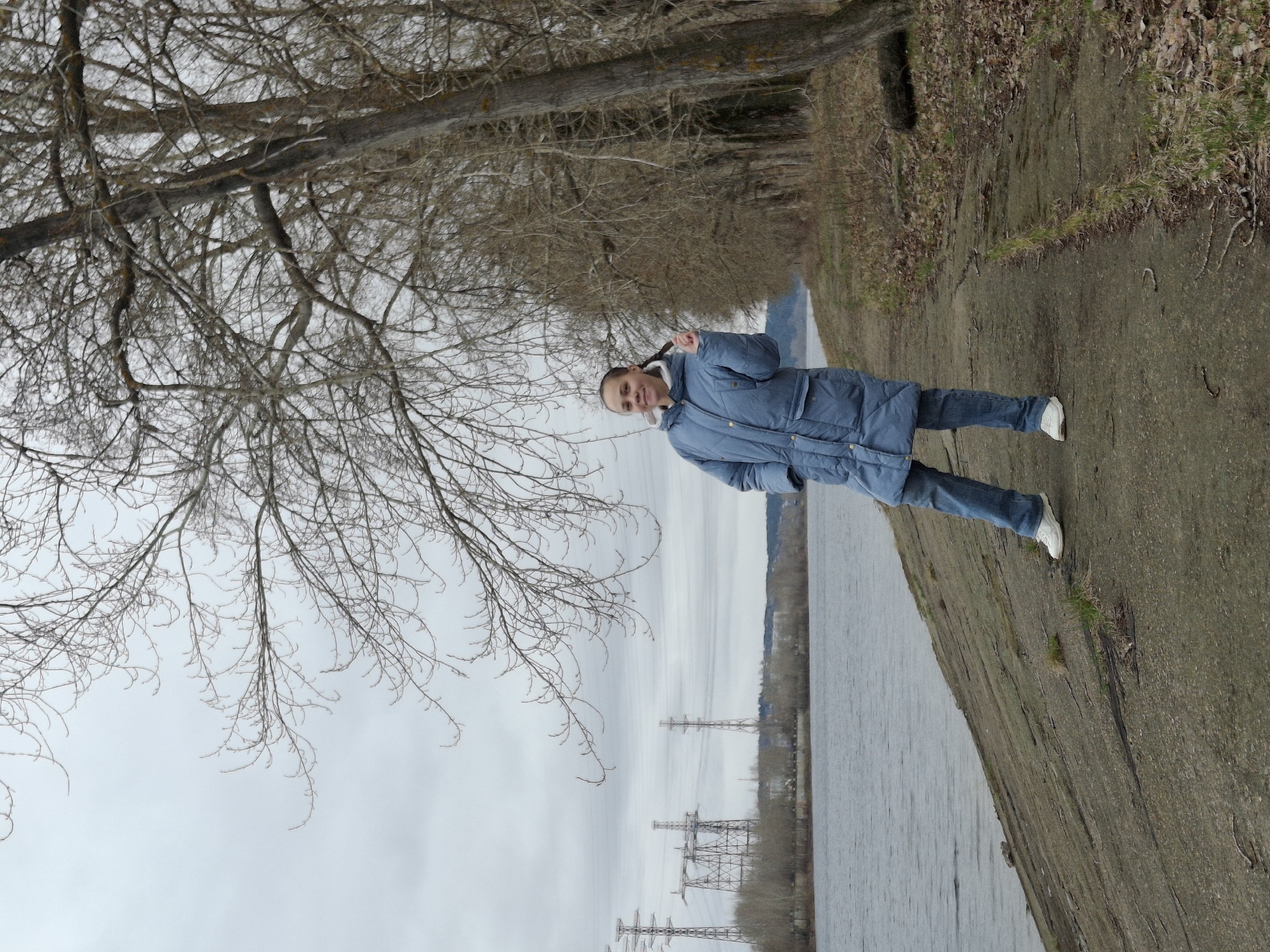This daytime image, captured in Chaikovsky, Russia, features a young woman posing cheerfully on a path beside a body of water, likely a river, under an overcast sky. The woman is the central subject, standing upright and smiling directly at the camera, her right hand raised in a peace sign. She is dressed in a long blue winter puffer coat, blue jeans, and white sneakers, with her dark hair pulled back.
To her right, a calm, wide expanse of water stretches into the distance, reflecting the muted tones of the grey sky. Across the water, several large electricity pylons are visible against a backdrop of distant land and bare trees, hinting at an industrial or developed landscape. To the woman's left, a dense array of leafless tree branches creates a natural canopy over the path, indicating a colder season such as late autumn or winter. The ground beneath her is a mix of dry earth and sparse, brown vegetation. The overall atmosphere suggests a brisk outdoor setting. There is no visible text in the image.