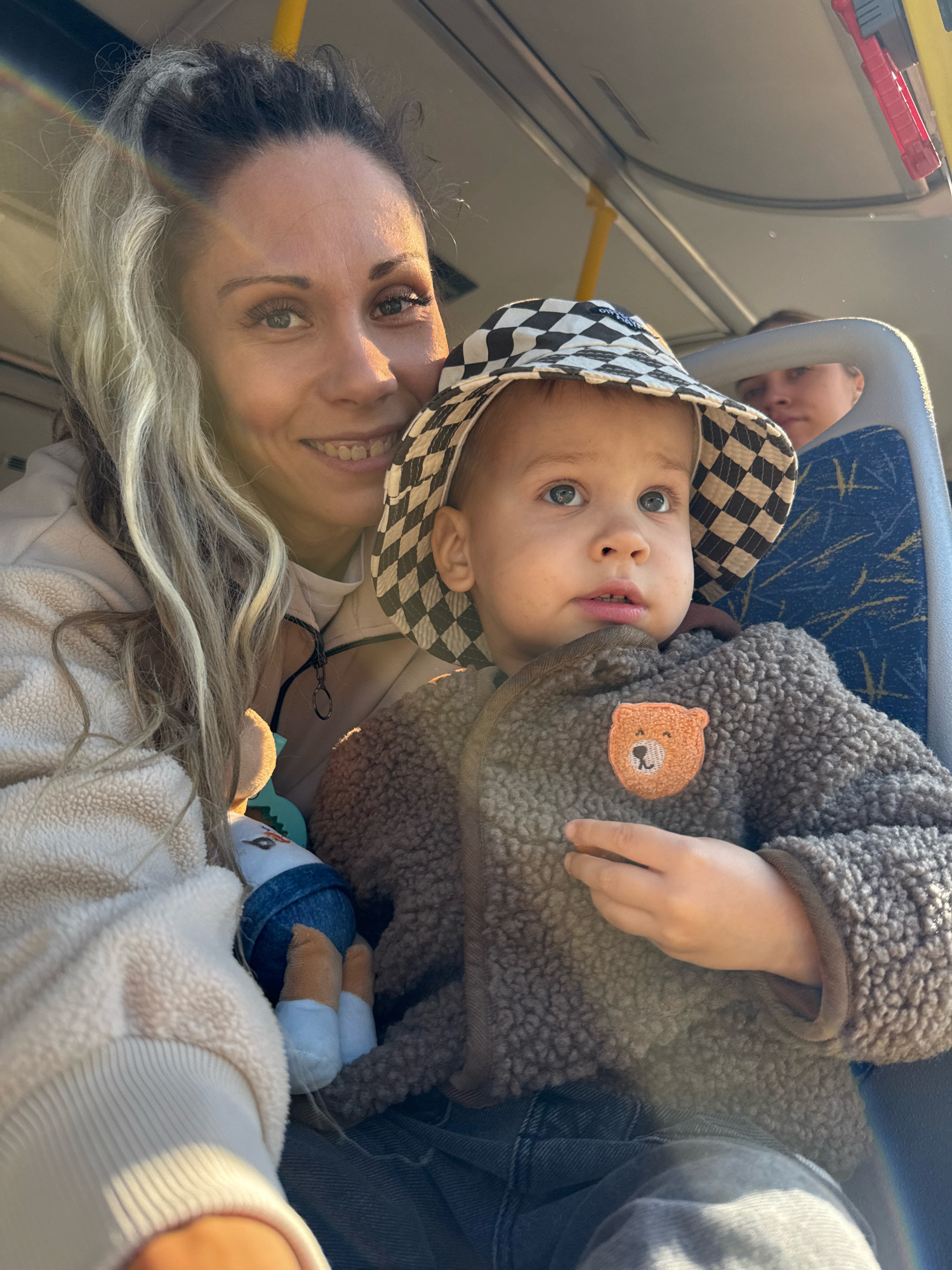 In this close-up, eye-level shot, a woman and a young child are seated on a bus, likely traveling within the city of Rusanovo, Russia. The woman, positioned on the left, is smiling and looking towards the camera. Her blonde hair, with streaks of grey, is pulled back. She is wearing a light-colored hooded sweatshirt. The child, a boy with blue eyes, is in the foreground, wearing a checkered hat and a fluffy brown jacket with a bear patch. He is looking upwards and slightly to the right. A plush toy is visible between the woman and the child. In the background, another person is partially visible seated further down the bus aisle. The interior of the bus is suggested by the yellow pole, grey ceiling, and a blue and grey patterned seat. Sunlight streams in from the side, casting a warm glow and creating lens flare effects on the woman's hair and the lower portion of the image.